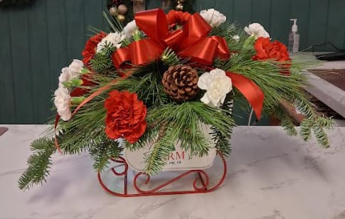 Winter Sleigh Ride - Gliding in with holiday charm, Winter Sleigh Ride is a festive Christmas arrangement designed in a beautifully detailed sleigh. Lush evergreens cradle rich red roses, cheerful carnations, snowy white blooms, and sparkling seasonal accents that capture the magic of winter. Textured holiday greens, berries, and subtle shimmer bring depth and elegance, making this piece feel warm, whimsical, and wonderfully nostalgic. A perfect centerpiece or gift to spread joy all season long.