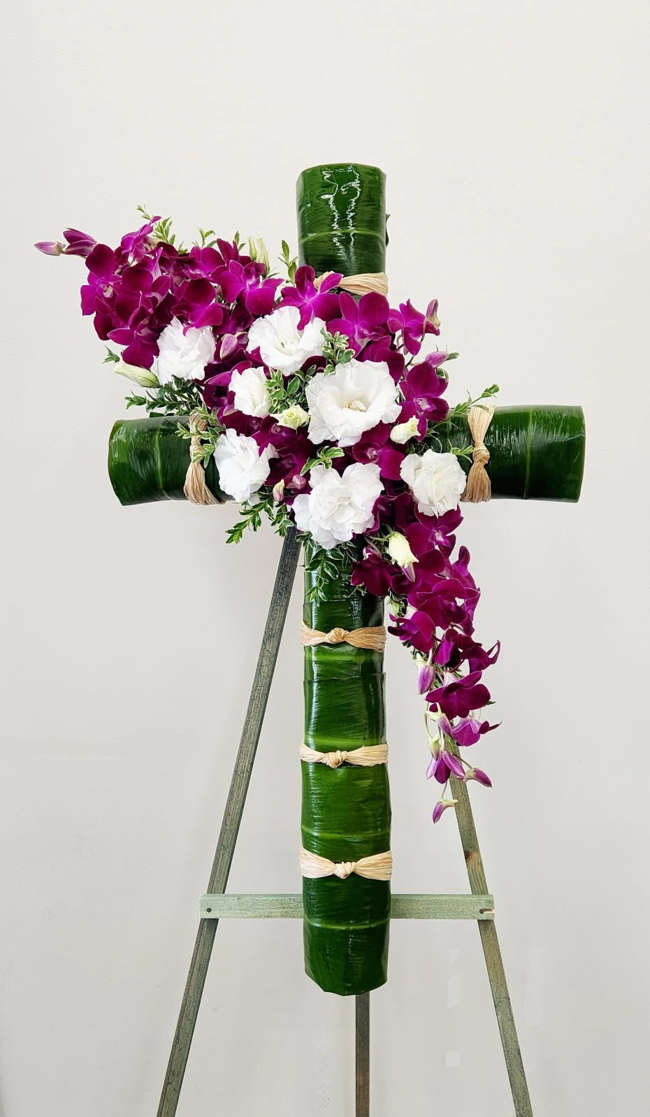Glorious Life Cross - Honor a life well-lived with this striking floral cross. Lush green ti leaves form the foundation, bound together with natural raffia for an earthy, organic touch. Purple dendrobium orchids cascade gracefully across the face, creating a stunning focal point that speaks to beauty, strength, and eternal remembrance. A meaningful tribute perfect for celebrations of life and cherished memories.  ONE SIZE ONLY 32"