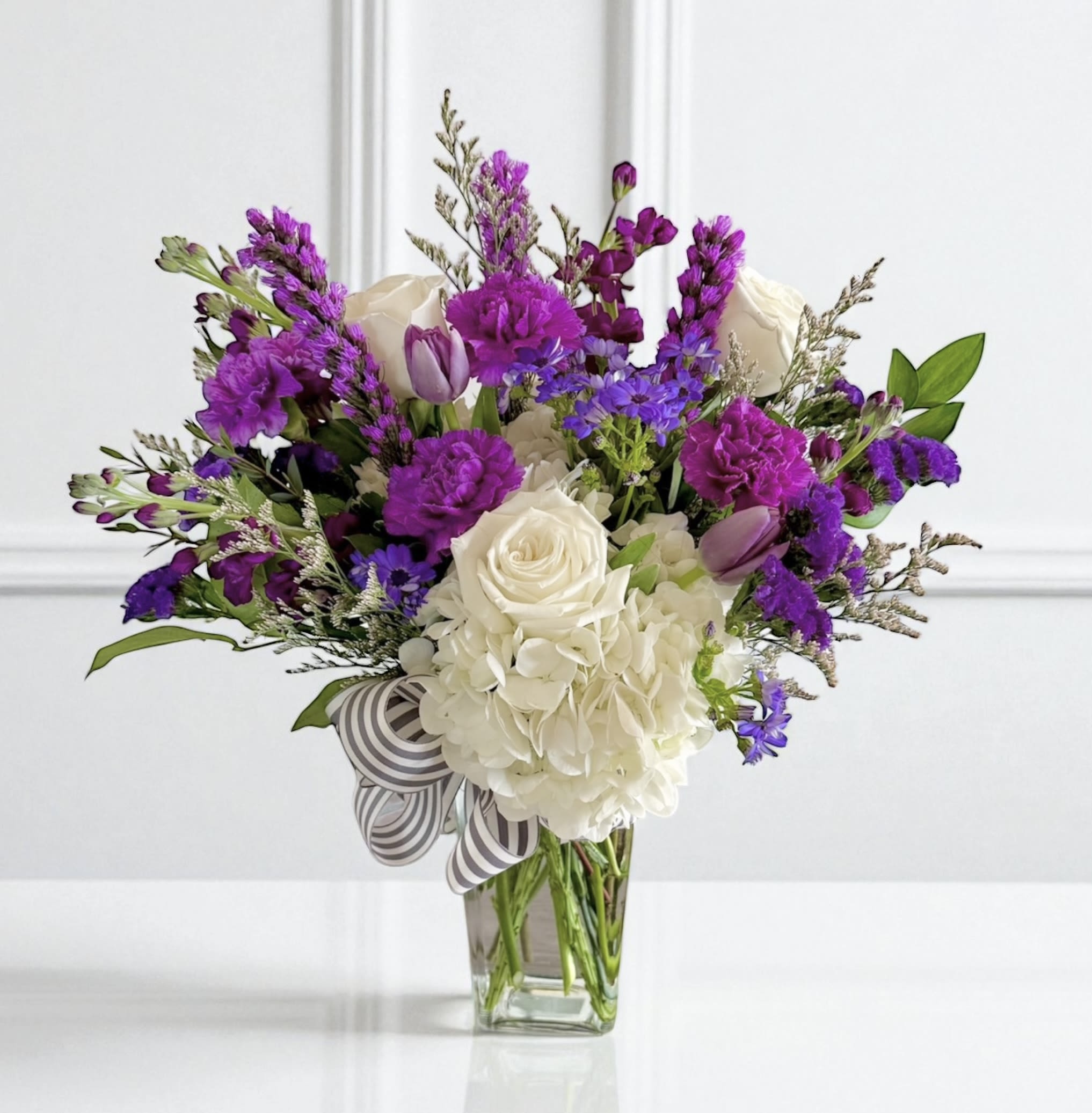 The Luka  - A taller clear vase filled with all purple and white flowers. Flowers may vary per season but will always be a purple and white mix. Included in the photo are hydrangeas, liatris, stock, roses, carnations, asters and statice!  