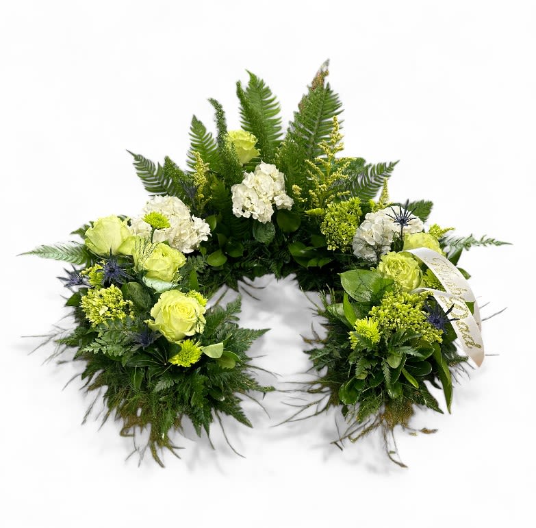 Tranquil Harmony Urn Surround - Tranquil Harmony is a serene and elegant funeral urn arrangement that encapsulates the beauty of nature and the essence of peace. This thoughtfully designed display features a lush variety of ferns that gracefully cascade around the base, symbolizing eternal life and renewal.   The arrangement highlights the gentle tones of white and green hydrangeas, their full blossoms exuding a sense of calm and comfort. The thistle's accents add texture and unique beauty, while the soft green roses bring a sense of hope and tranquility. Nestled among these elements, cheerful green mums provide a vibrant contrast, representing cherished memories.  Together, these elements create a visual serenade that honors the life of a loved one, inviting those who view it to reflect on the beauty of their journey while finding solace in the harmonious blend of colors and textures. Tranquil Harmony serves as a touching tribute that embodies both remembrance and peace.  Inside dimensions are 16&quot; in diameter