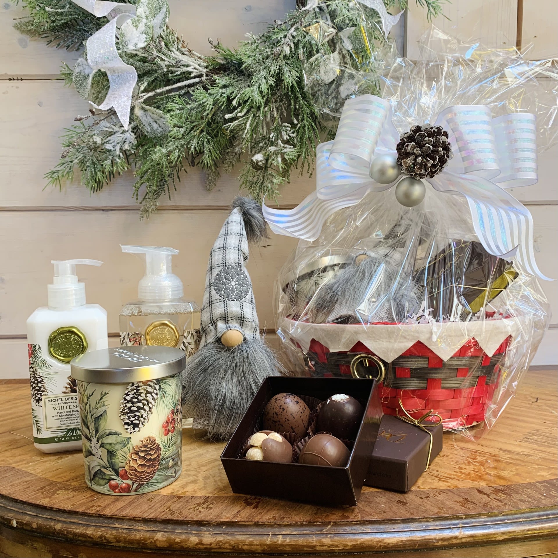 White Spruce Deluxe Gift Basket - This deluxe White Spruce gift basket includes a gnome, a four piece box of chocolates, one hand and body lotion, one foaming hand soap, and one scented decorative glass candle beautifully wrapped up and ready to go with a large bow and pinecones with ornament accent. Fragrance: True Spruce with touches of fruit and spice  1) A hand picked just for you Christmas gnome with an $11.95 value  2) A 4-piece box of our specialty chocolate truffles made in the USA from imported Belgian chocolate. Pictured flavors are mocha, macadamia hazelnut, milk chocolate and coconut, substitutions available on request, or occasionally necessary due to supply. Contains nuts   3) Our hand and body lotion is made with shea butter, aloe, and other botanical ingredients and packaged in a decorative pump bottle. 12 fl. oz. / 354 ml lotion. 4) Our foaming hand soap contains luxurious shea butter and aloe vera for gentle cleansing and moisturizing. 17.8 fl. oz. / 530 ml liquid  5) Our all-natural, decorative glass candle is nontoxic, biodegradable and clean burning, made from a sustainably sourced soy and palm oil wax blend. This beautiful glass jar with lid can be used at home, indoors or out. 7.4 oz./209 g. Over 30 hrs. approximate burn time.  Customizable gift baskets available upon request. Please give us a call at 775-348-6161 for assistance putting together the perfect aromatherapy basket for your loved one. 