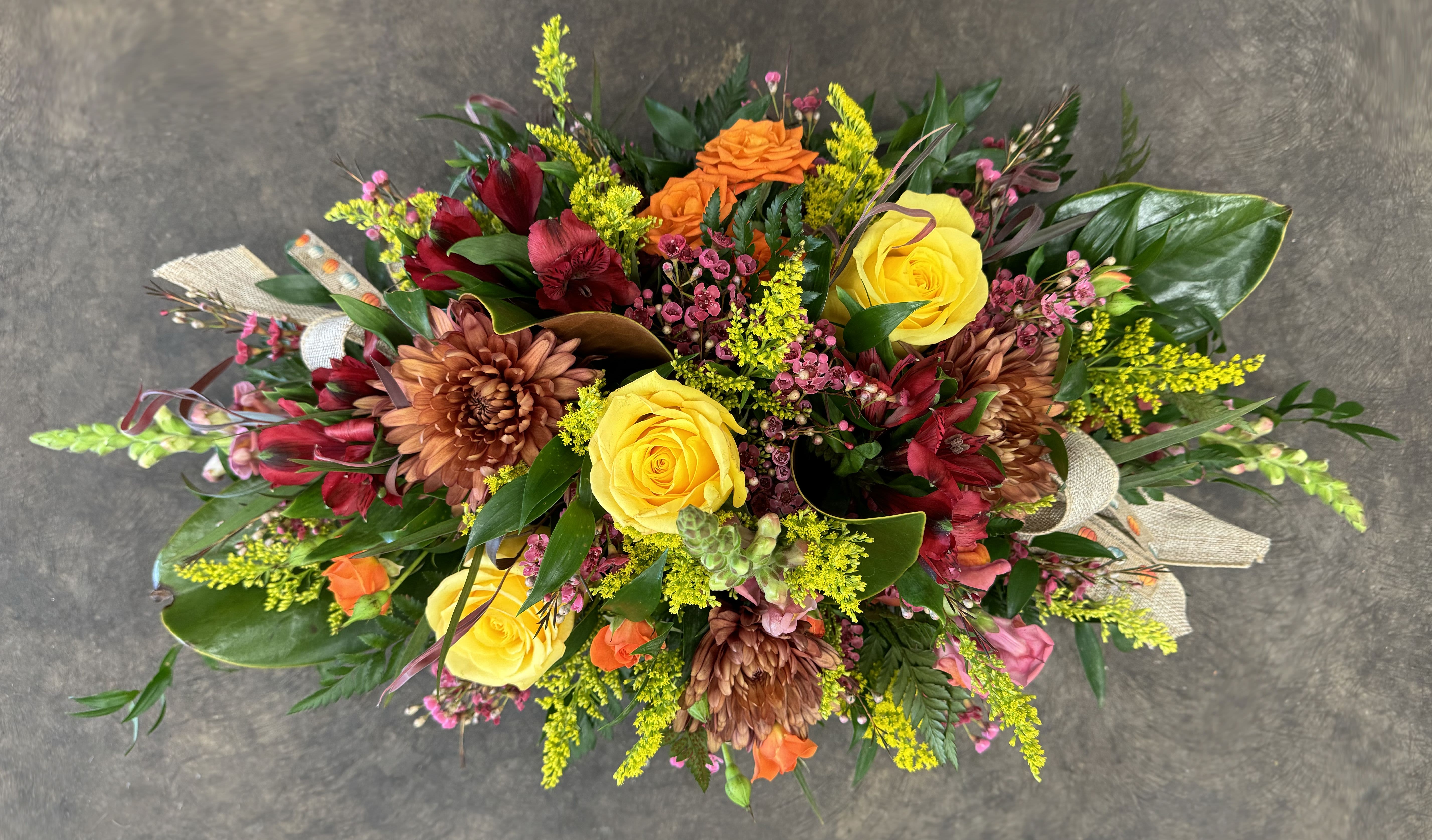 Thanksgiving Abundance Centerpiece Basket - Embrace the spirit of gratitude and harvest with our Thanksgiving Abundance Basket, a stunning floral arrangement designed to enhance your holiday table or serve as a meaningful gift. Crafted in a rustic wicker basket, this luxurious piece features a radiant blend of golden yellow roses, orange spray roses, and deep burgundy alstroemeria, complemented by waxflower, solidago, and lush greenery. Perfect as a centerpiece for Thanksgiving gatherings or as a beautiful gesture of appreciation, this arrangement captures the essence of the season’s abundance and warmth. Whether gracing a dinner table or expressing heartfelt thanks to someone special, this basket brings both beauty and sentiment to any occasion. 