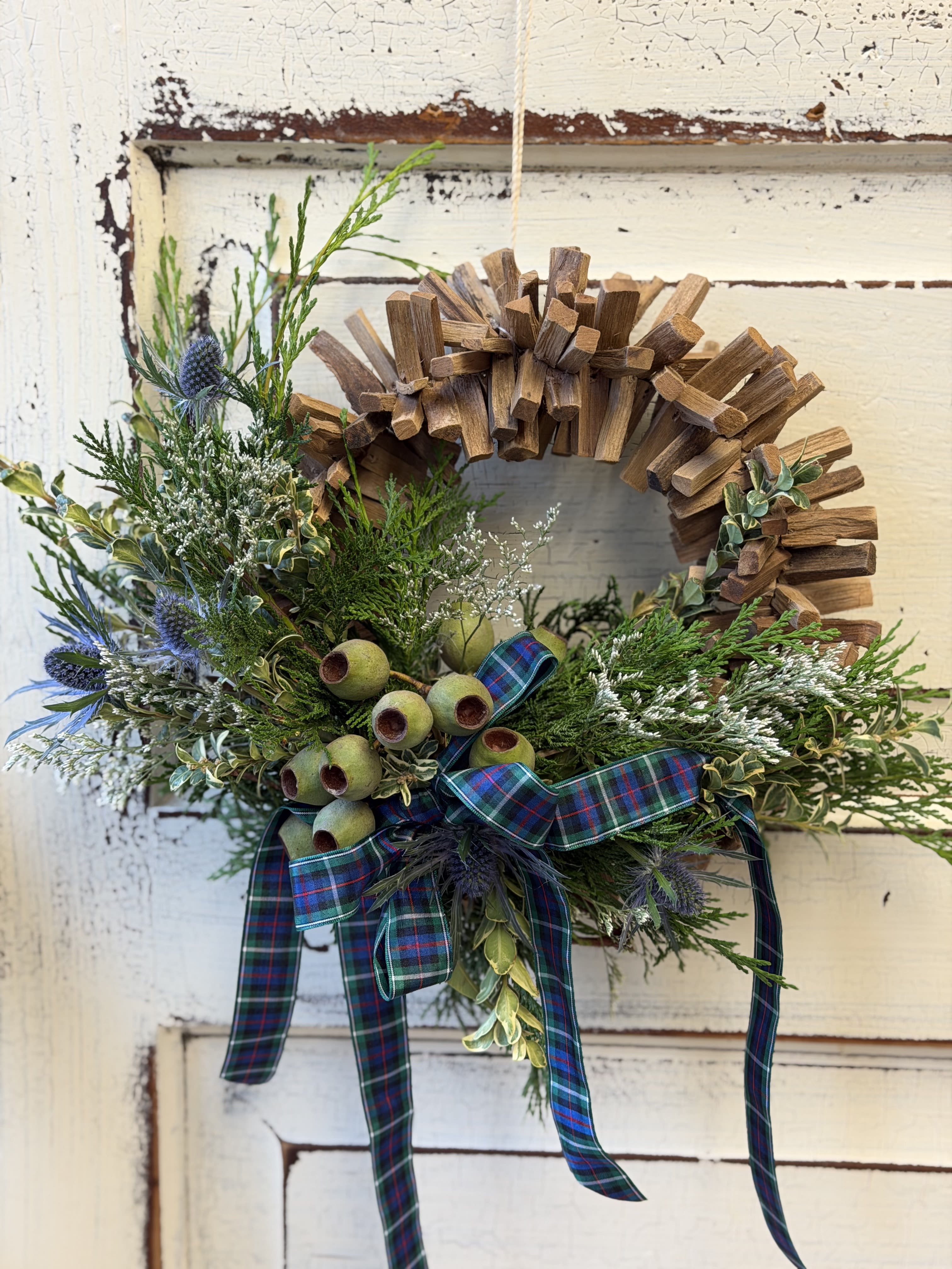 Mini Winter Wreath - A little modern, a little rustic, this wreath has the scents of the season. The base is a unique driftwood construction that looks great on its own. Fresh evergreens, dried flowers and pods, and ribbon complete the look. Wreath is about 12&quot; and arrives ready to either hang or place on the table as a candle ring.