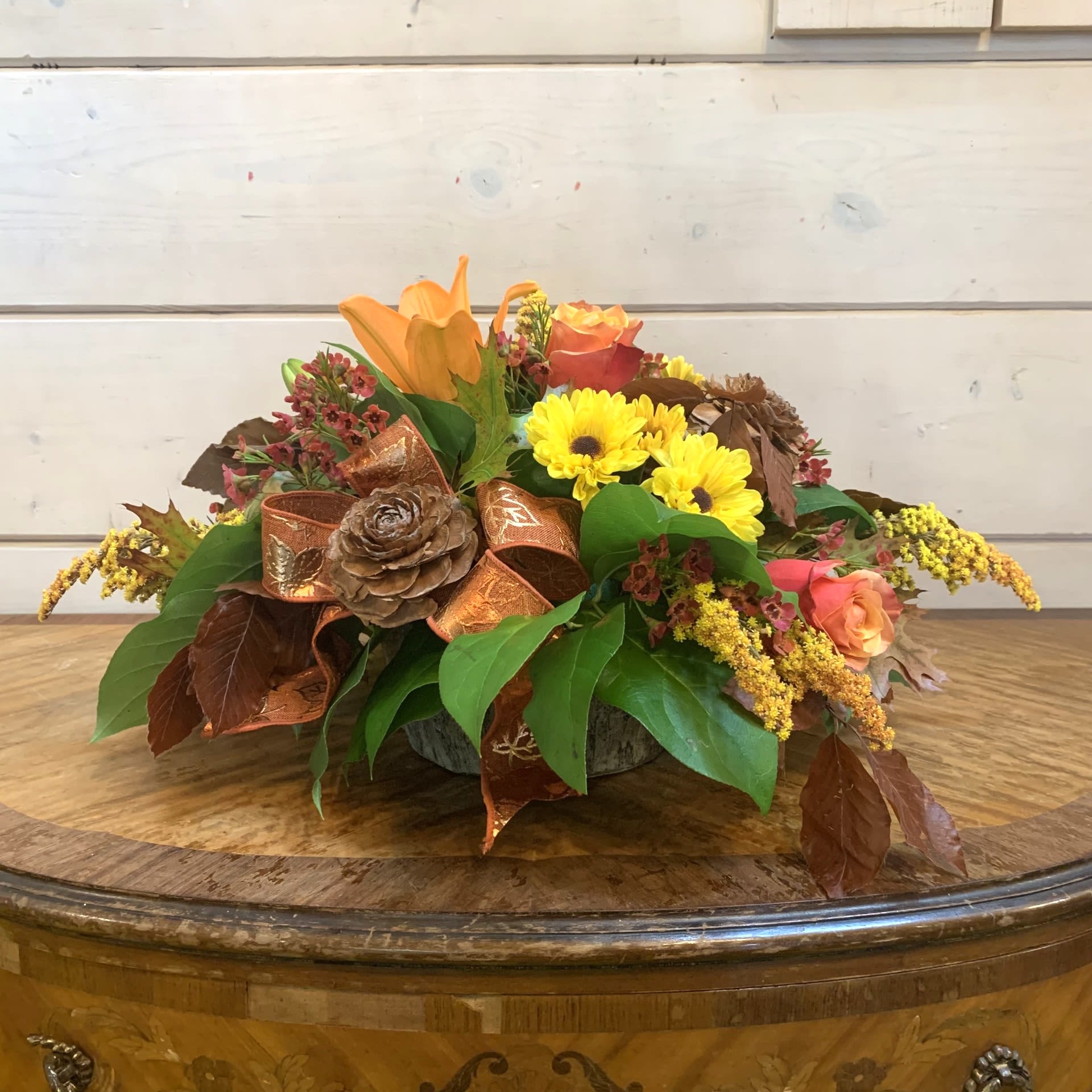 Grateful Gatherings Centerpiece - Gather around the table with this arrangement of orange roses, viking yellow daisies, and orange lilies in a low bark pot with seasonal fall foliage, fall solidago, fall wax flowers, rosette pinecones, and bronze maple leaf bows. Approximately 13&quot; across for the standard, 14&quot; for the deluxe and 15&quot; for the premium.