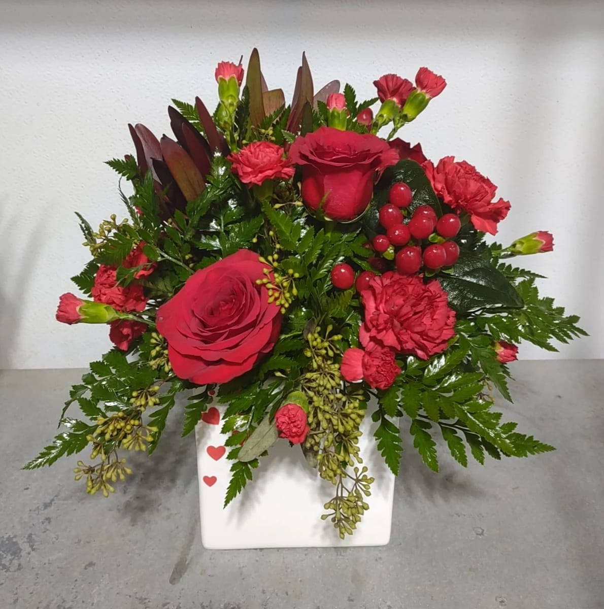 Fluttering hearts - A modern twist on a tradtional favorite.  A white square container with little red hearts on the side featuring roses, safari sunset, mini carnations and berries. 