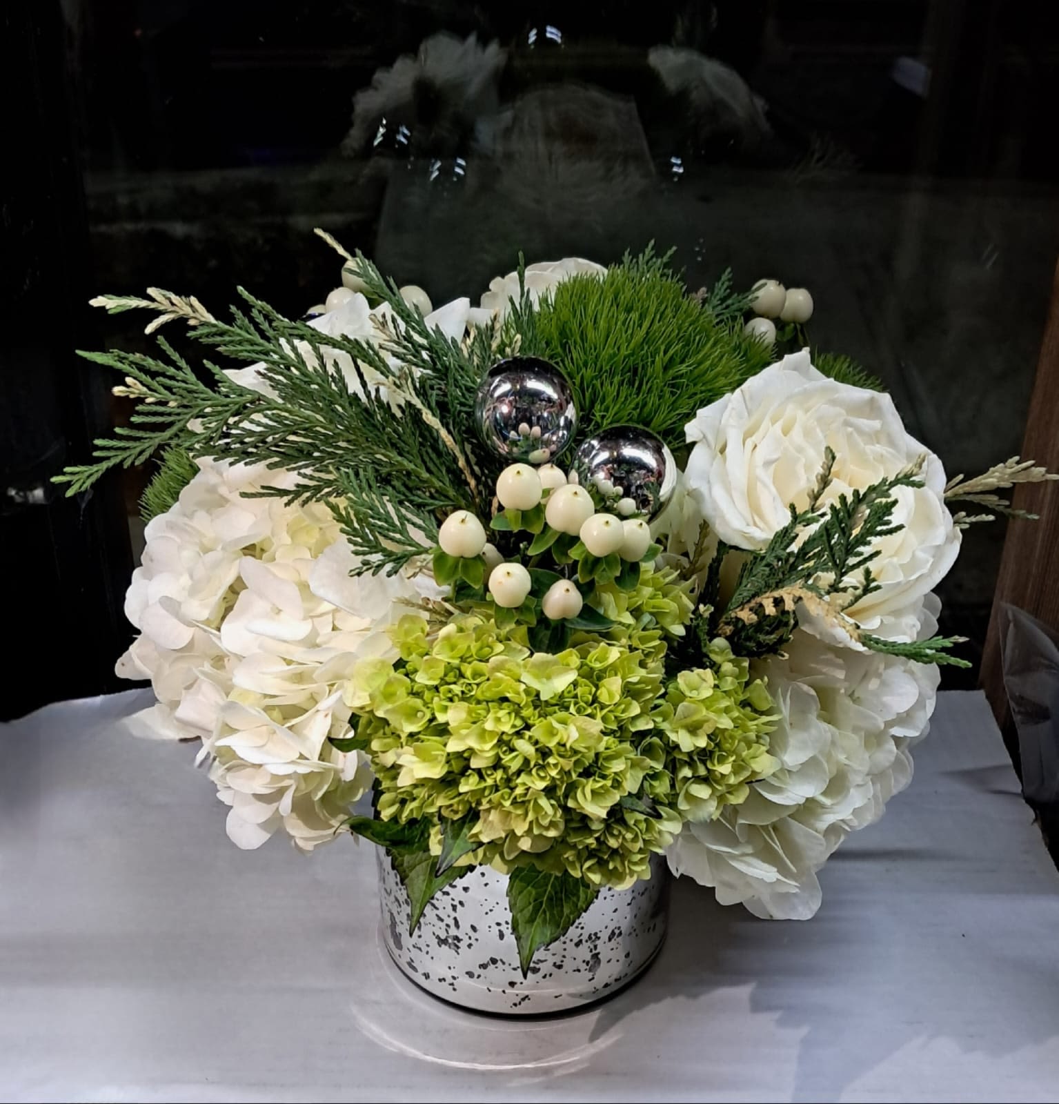 Silver Skies  - This beautiful arrangement has white roses, white and green hydrangeas, hypericums, small ornamental spheres and greenery. It come in a silver vase. 