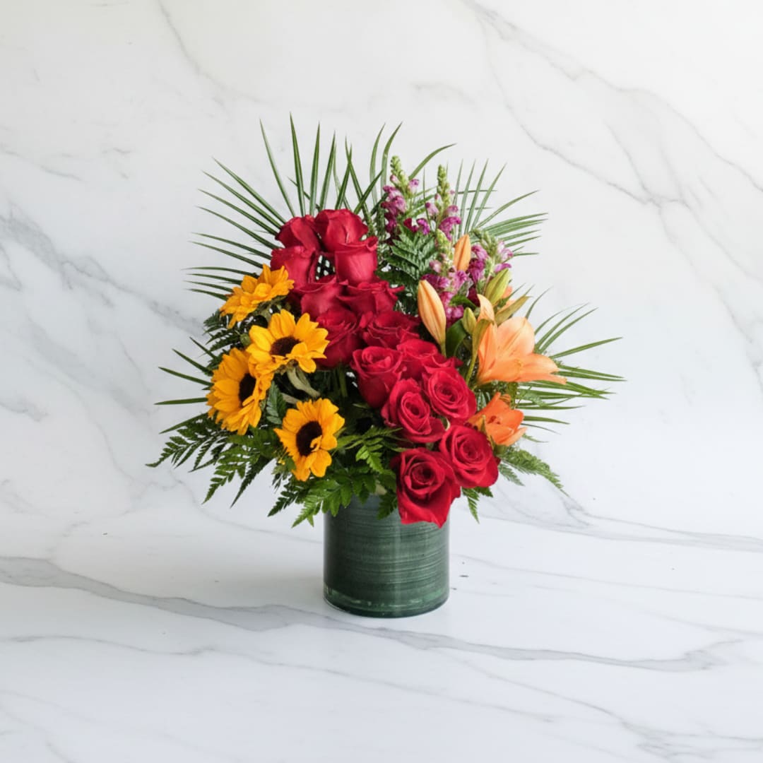 Sunset Radiance - A joyful celebration of color and elegance. This handcrafted arrangement features vibrant red roses, sunny mini sunflowers, soft orange lilies, and delicate snapdragons, all designed beautifully with lush tropical greenery. Perfect for birthdays, congratulations, or simply bringing warmth into any room, this arrangement blends premium blooms with a modern, elevated style.