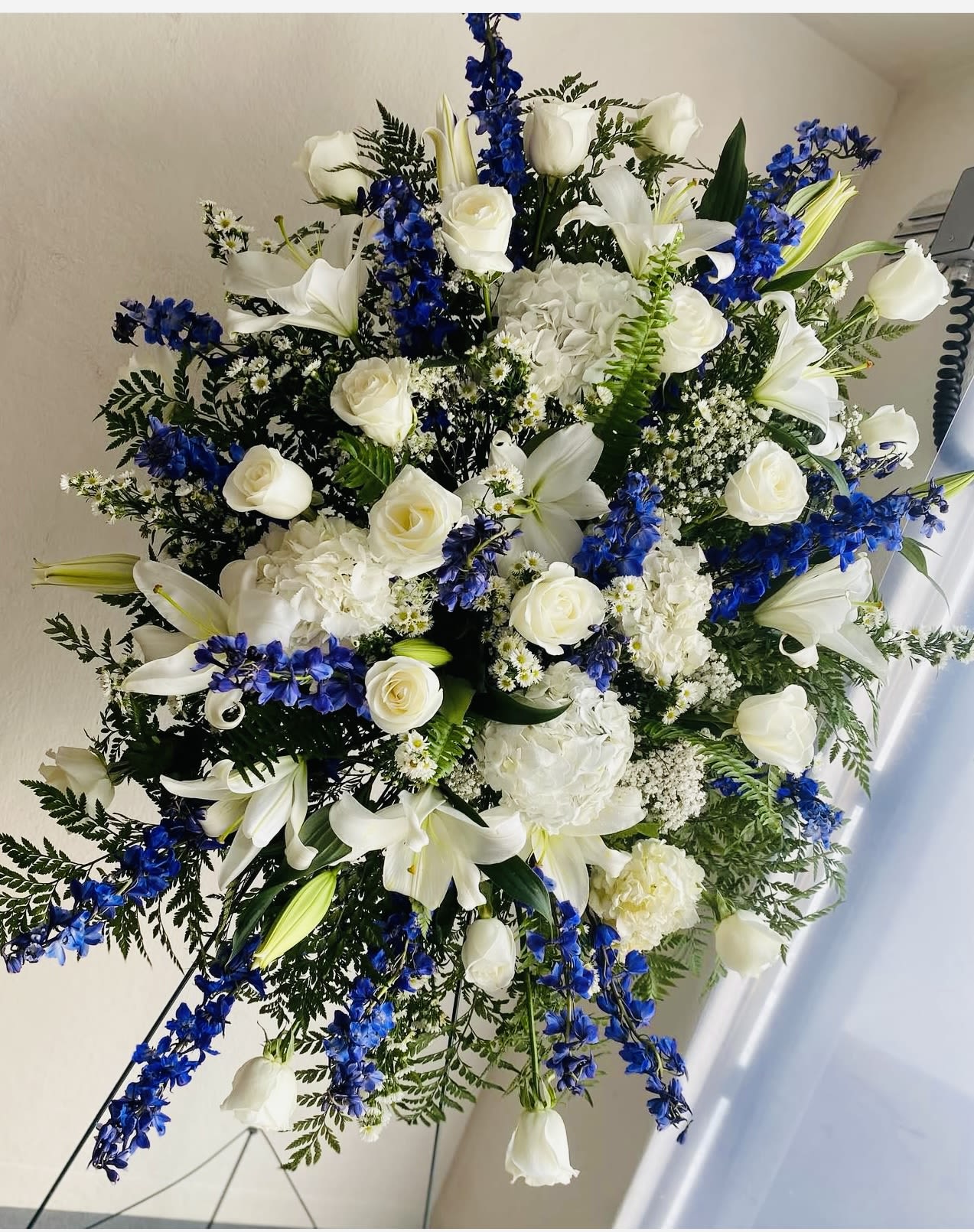 White and blue standing spray easel  - A serene and elegant tribute, this White and Blue Standing Spray beautifully conveys peace, dignity, and heartfelt remembrance. Expertly arranged on a standing easel, this graceful display features a soothing palette of crisp white blooms and cool blue accents, symbolizing purity, tranquility, and everlasting love.  The design often includes white lilies, roses, chrysanthemums, and carnations, complemented by blue delphiniums or hydrangeas for a touch of gentle color. Lush greenery—such as leatherleaf fern, eucalyptus, and salal—adds depth and texture, enhancing the natural beauty of the arrangement.