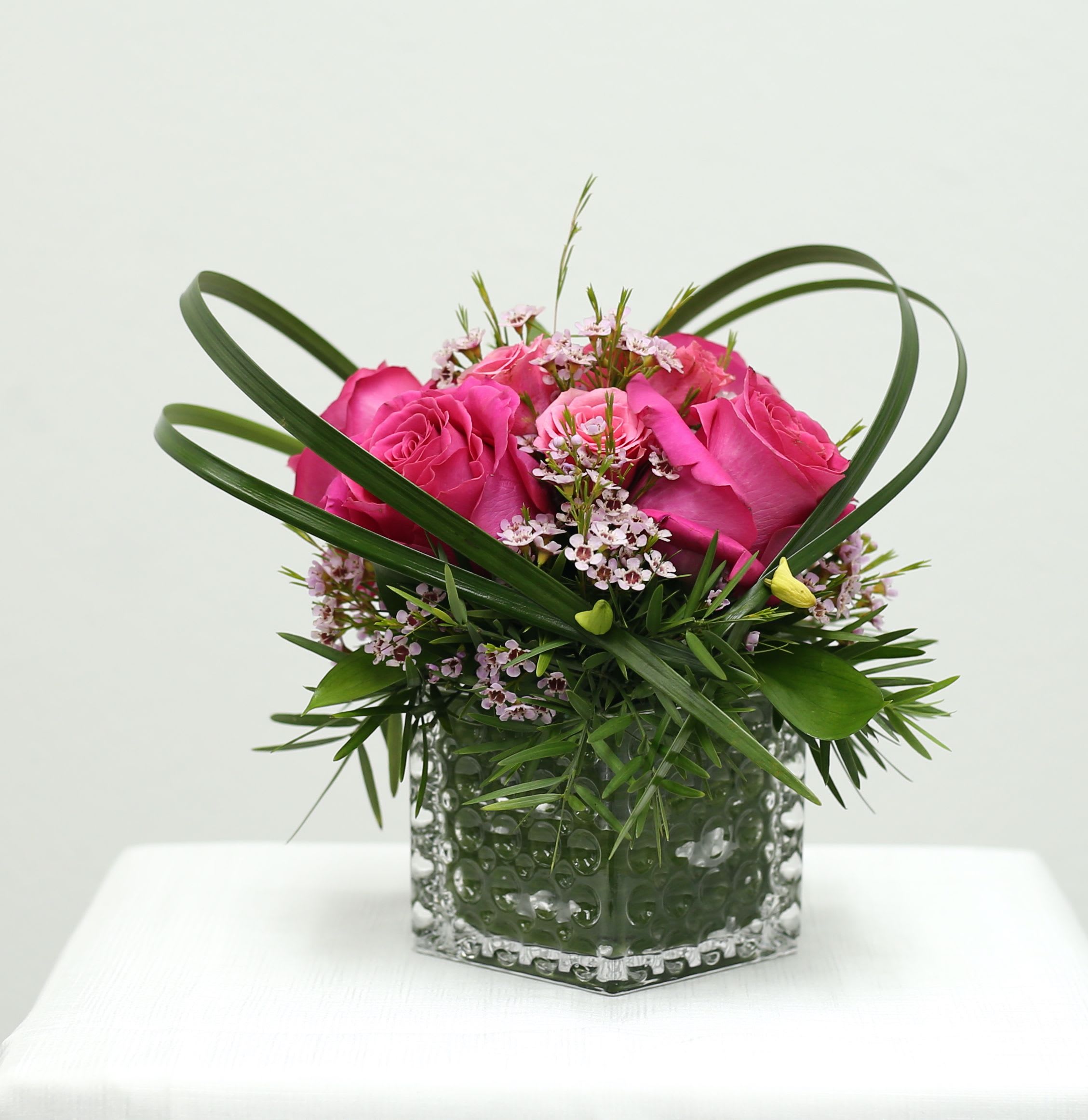 Rose Jewel - Bathed in soft pink hues, this exquisite arrangement features lush roses nestled amongst delicate blooms. The interplay of light and shadow dances across the textured glass vase, enhancing the ethereal beauty of the blossoms within. A touch of refined elegance for any cherished space.