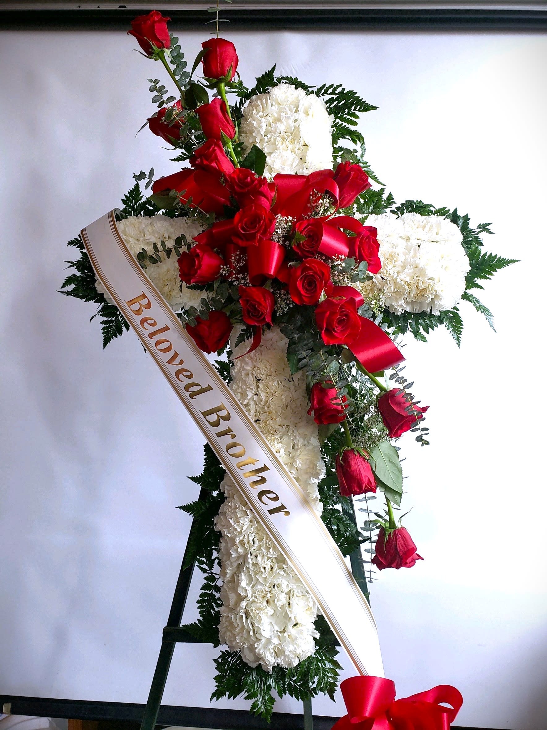 Sympathy Cross #42 - All white standing cross with a cluster of red roses. Colors can be modified depending on availability. Designed on an easel that is included with the arrangement and does NOT need to be returned. Banner is included with arrangement. Please specify what is to be printed on banner upon check out under "FLORIST INSTRUCTIONS". LIMITED TO 5 WORDS ON BANNER.