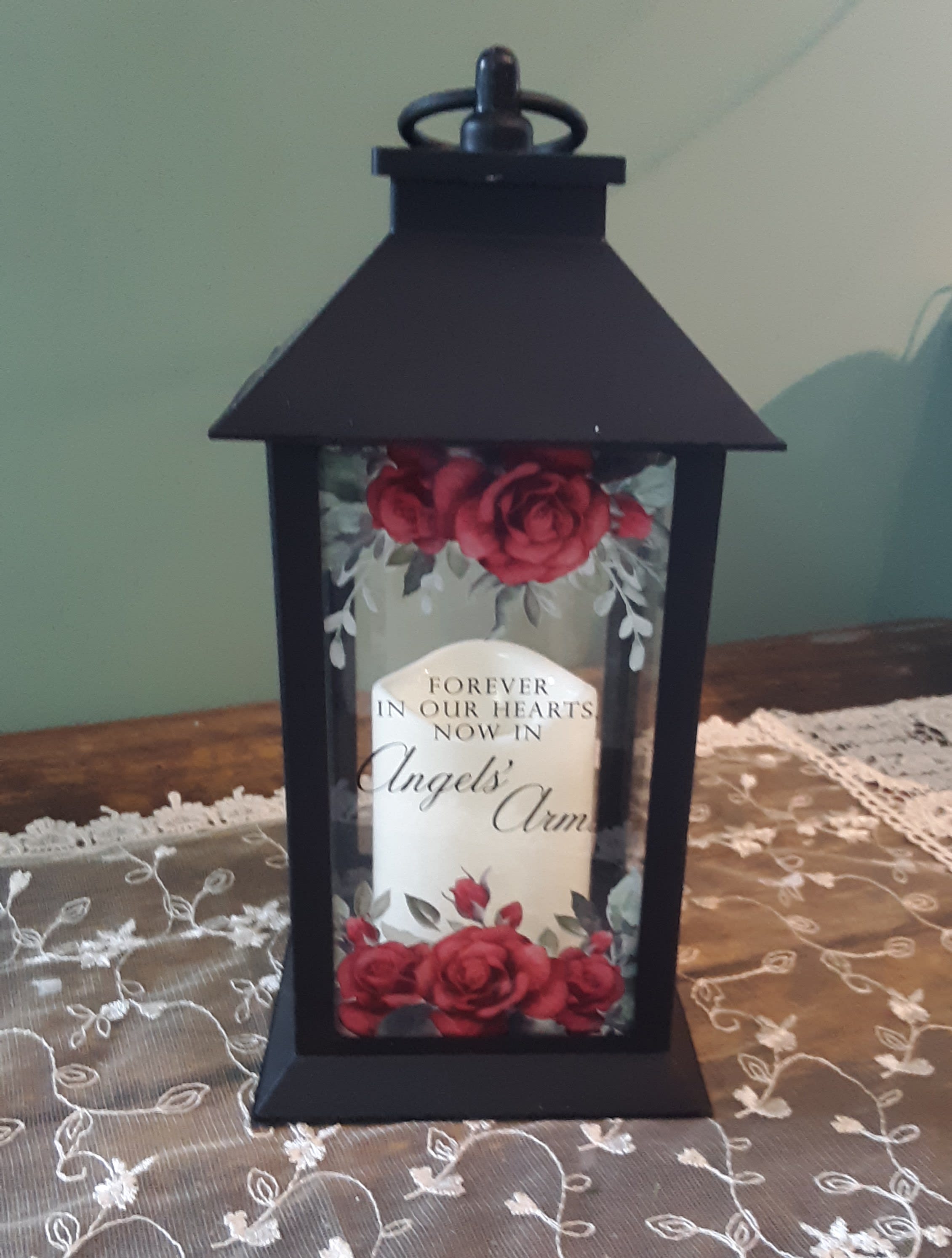 "Angels' Arms Rose Lantern" - This beautiful black high quality polyresin finish lantern surrounds an interior of a flameless LED candle. On the front panel are clusters of classic red roses as well as the saying "FOREVER IN OUR HEARTS, NOW IN ANGELS' ARMS".    Lantern is 13"H x 5 1/2"W x 5 1/2" D.  A built in automatic timer and 3 AAA batteries are included.    This item is suitable for sympathy gifting either at the vistation or the home and is weather friendly.  