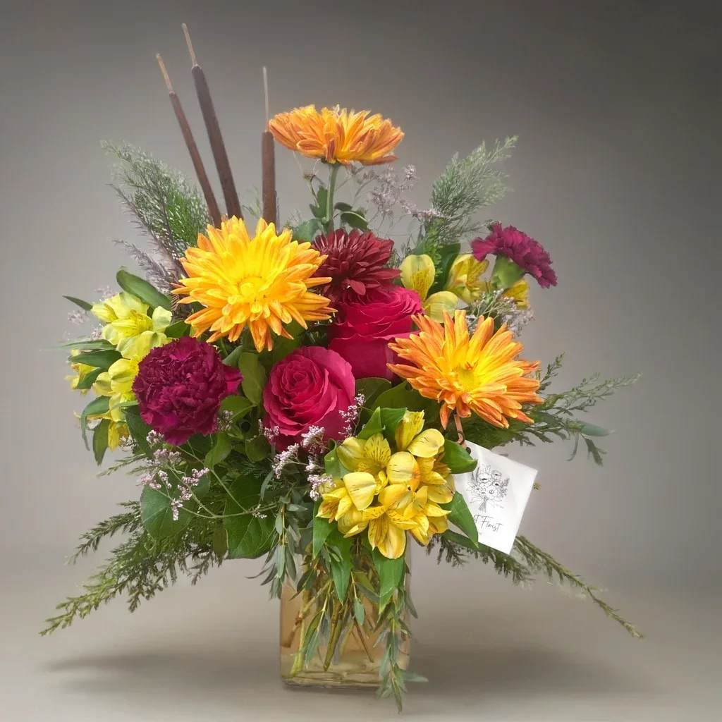 Golden Harvest - Bring the warmth of the season to your home with our Golden Harvest Thanksgiving Centerpiece, a lush fall flower arrangement designed to brighten your holiday table. This autumn bouquet features vibrant orange and yellow mums, deep cranberry blooms, fresh roses, yellow alstroemeria, and seasonal greenery arranged in a modern vase for a full, abundant look.  Perfect for Thanksgiving dinner, Friendsgiving, or fall gatherings, this handcrafted arrangement adds rich color and festive charm to any space. It also makes a thoughtful hostess gift and is available for fast, reliable local delivery.  If you’re searching for Thanksgiving flowers, a fall floral centerpiece, or the perfect autumn table arrangement, this design brings all the beauty of the season in one elegant display.  Approximately 14&quot; x 18&quot;  Available for delivery in the Quad Cities and surrounding area.
