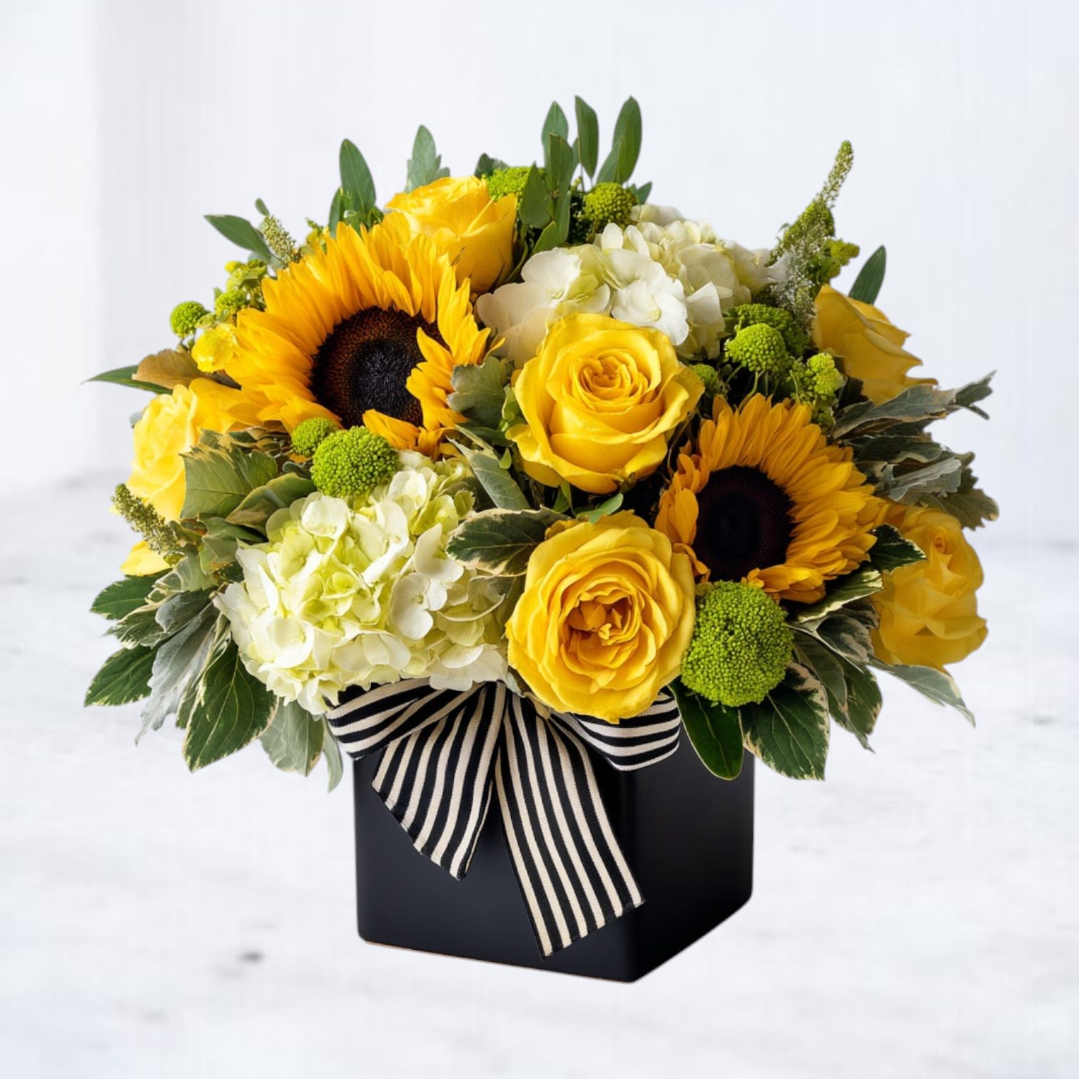 Solaris Charm - Radiate warmth and joy with the Solaris Charm arrangement. This vibrant design features golden sunflowers, bright yellow roses, delicate white hydrangeas, and green hypericum berries, artfully arranged in a sleek black vase and accented with a bold striped ribbon. Perfect for celebrating milestones, expressing gratitude, or simply bringing a burst of happiness to any space.  Why You'll Love It: Showcases a striking mix of sunny blooms for a bold statement. Ideal for birthdays, congratulations, or just-because moments. Modern black vase with a chic ribbon for a contemporary touch.