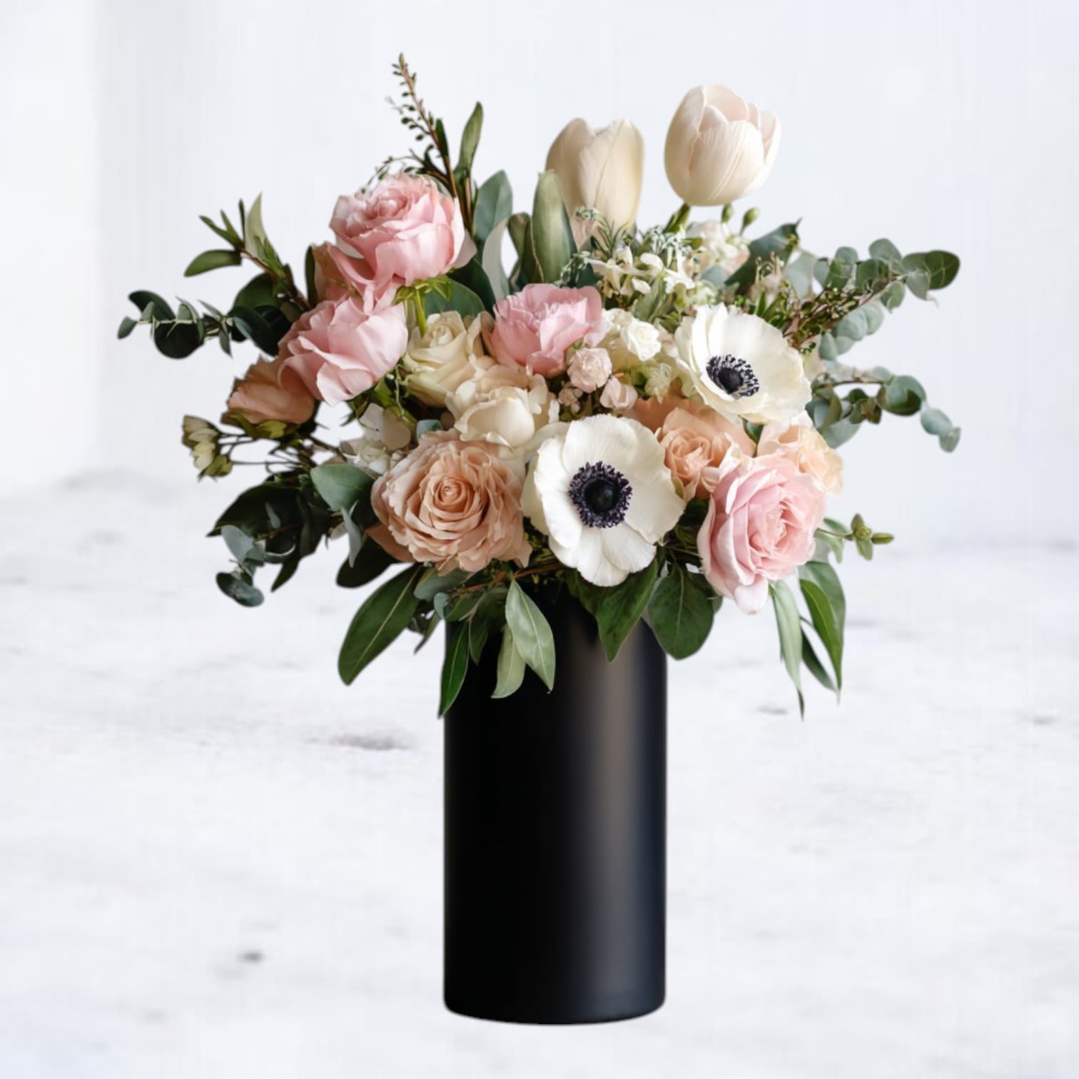 Noir Bloomscape - Elegant, modern, and effortlessly chic, Noir Bloomscape is a striking floral arrangement that blends soft romance with bold sophistication. Designed in an 8in sleek black vase, this arrangement features delicate blush roses, ruffled lisianthus, crisp white anemones, and fresh greenery, creating a timeless balance of texture and beauty.  Perfect for anniversaries, celebrations, or simply as an everyday statement piece, Noir Bloomscape exudes luxury and refinement. Available for same-day delivery in Houston and surrounding areas, ensuring your thoughtful gesture is delivered with elegance.
