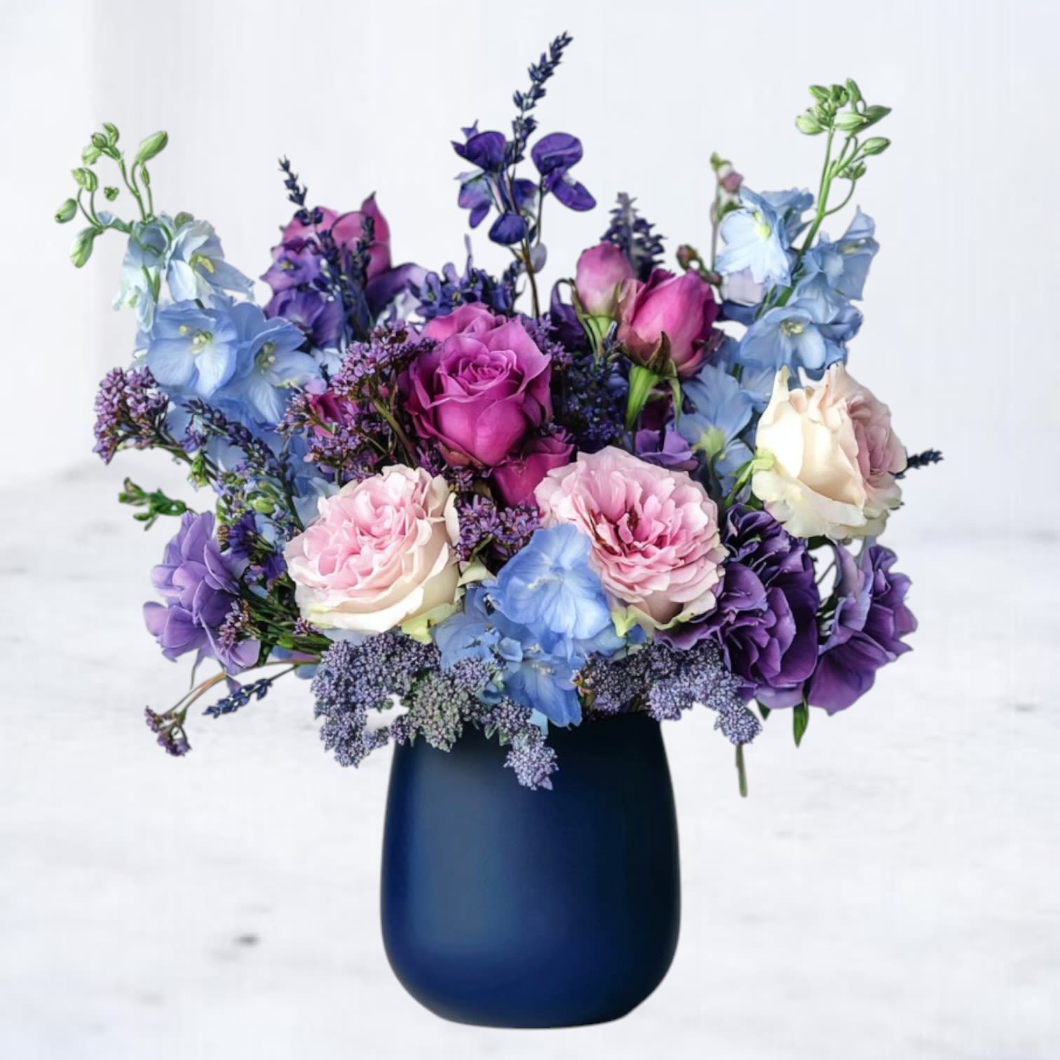 Dusk Reverie Bouquet - The Dusk Reverie Bouquet is a graceful fusion of soft blush and lavender roses, vibrant magenta blooms, and delicate blue delphiniums, artfully arranged in a sleek navy vase. This arrangement evokes the tranquil beauty of an evening sky, offering a timeless and elegant expression for anniversaries, or heartfelt moments.  Why You'll Love It: A romantic palette of deep, moody hues and soft pastels. Perfect as a sophisticated gift or a stunning home centerpiece. Hand-delivered with care for an unforgettable presentation. 