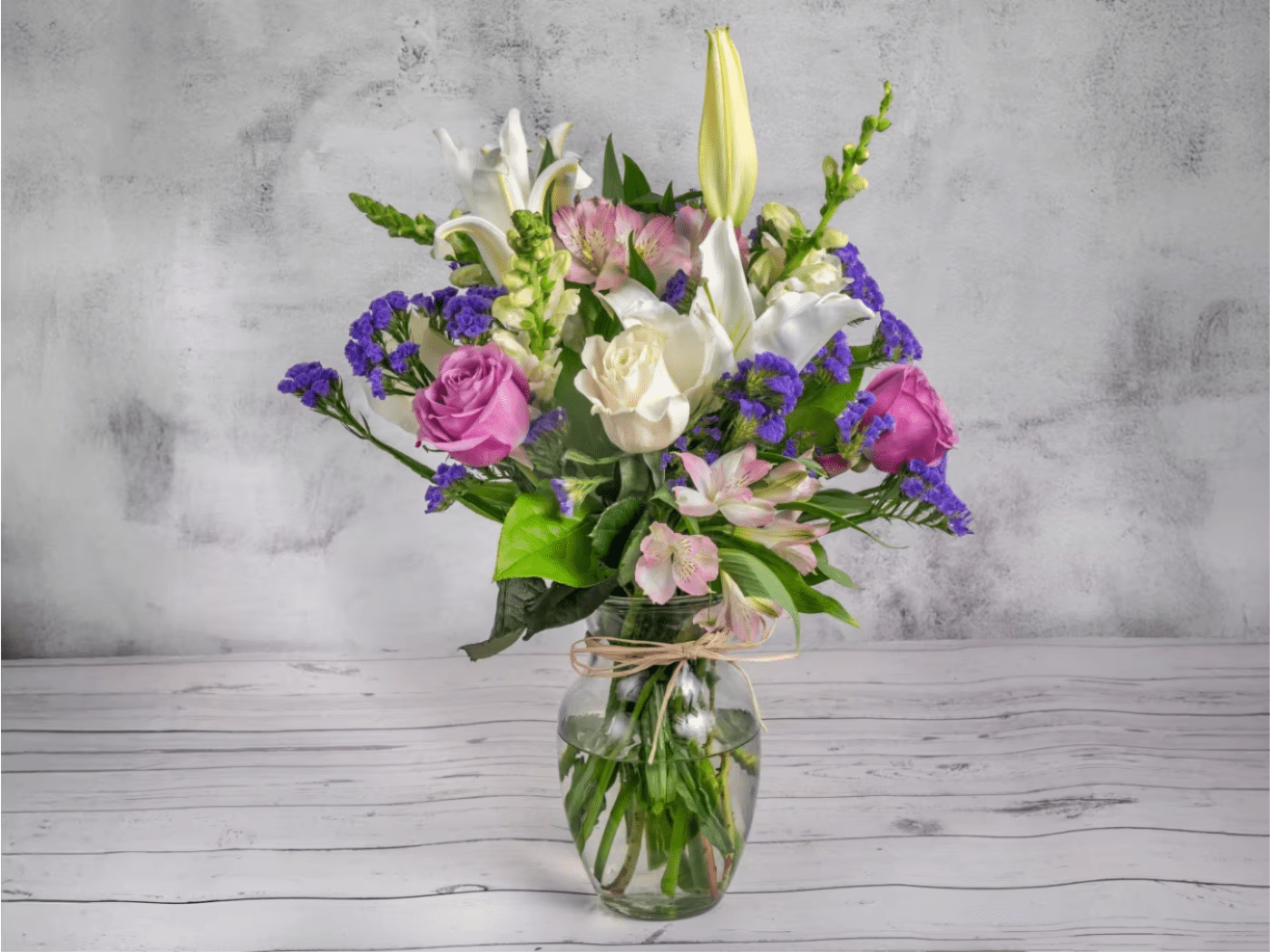 Pastel Whispers - A serene blend of white lilies, soft pink alstroemerias, lavender roses, and ivory blooms, highlighted with sprigs of deep purple statice. Arranged in a clear glass vase tied with natural raffia, this bouquet is a perfect symbol of elegance and warmth. Ideal for expressing heartfelt sentiments or adding a touch of grace to any setting.