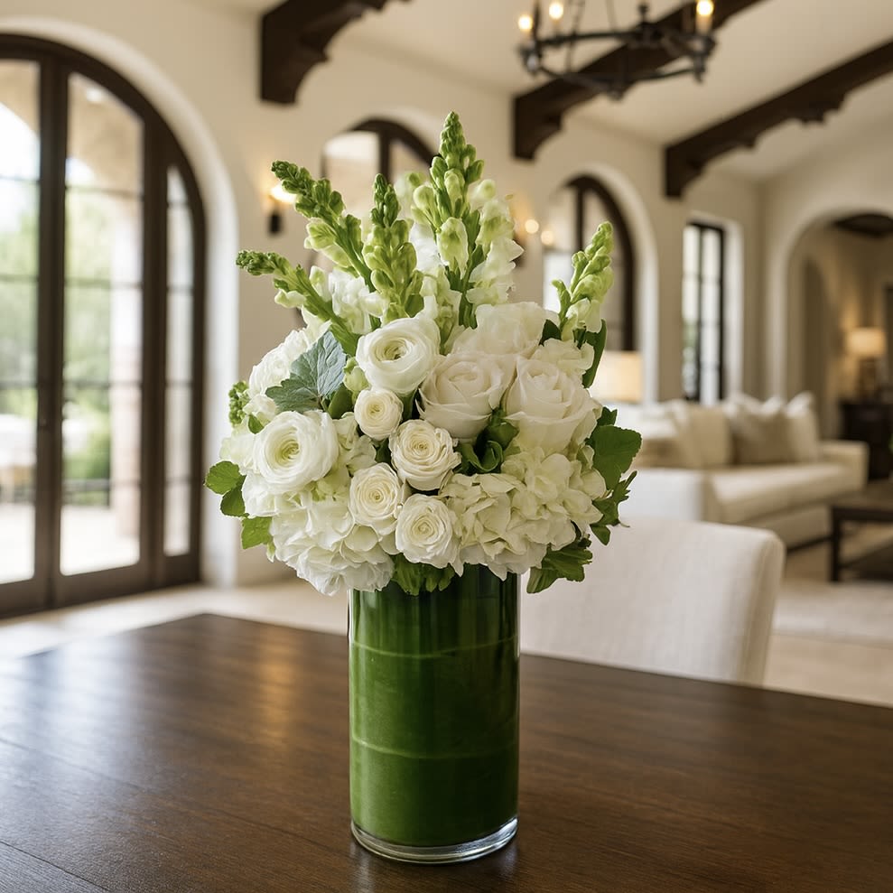 Angelique - Elevate any space with our Angelique Arrangement, a timeless blend of elegant white blooms designed to bring natural serenity and sophisticated beauty into your home. This luxurious centerpiece features premium white hydrangeas, garden roses, spray roses, ranunculus, and snapdragons, artfully gathered into a lush, full silhouette. Displayed in a modern glass vase wrapped with fresh green leaves, this arrangement offers a clean, organic aesthetic that complements any décor style.  Perfect for dining rooms, entry tables, kitchen islands, special events, anniversaries, sympathy gifts, or everyday home elegance, this all-white bouquet adds a soft, inviting glow to any interior.  Crafted by hand using the freshest seasonal stems, the Angelique Arrangement delivers long-lasting beauty and effortless sophistication.