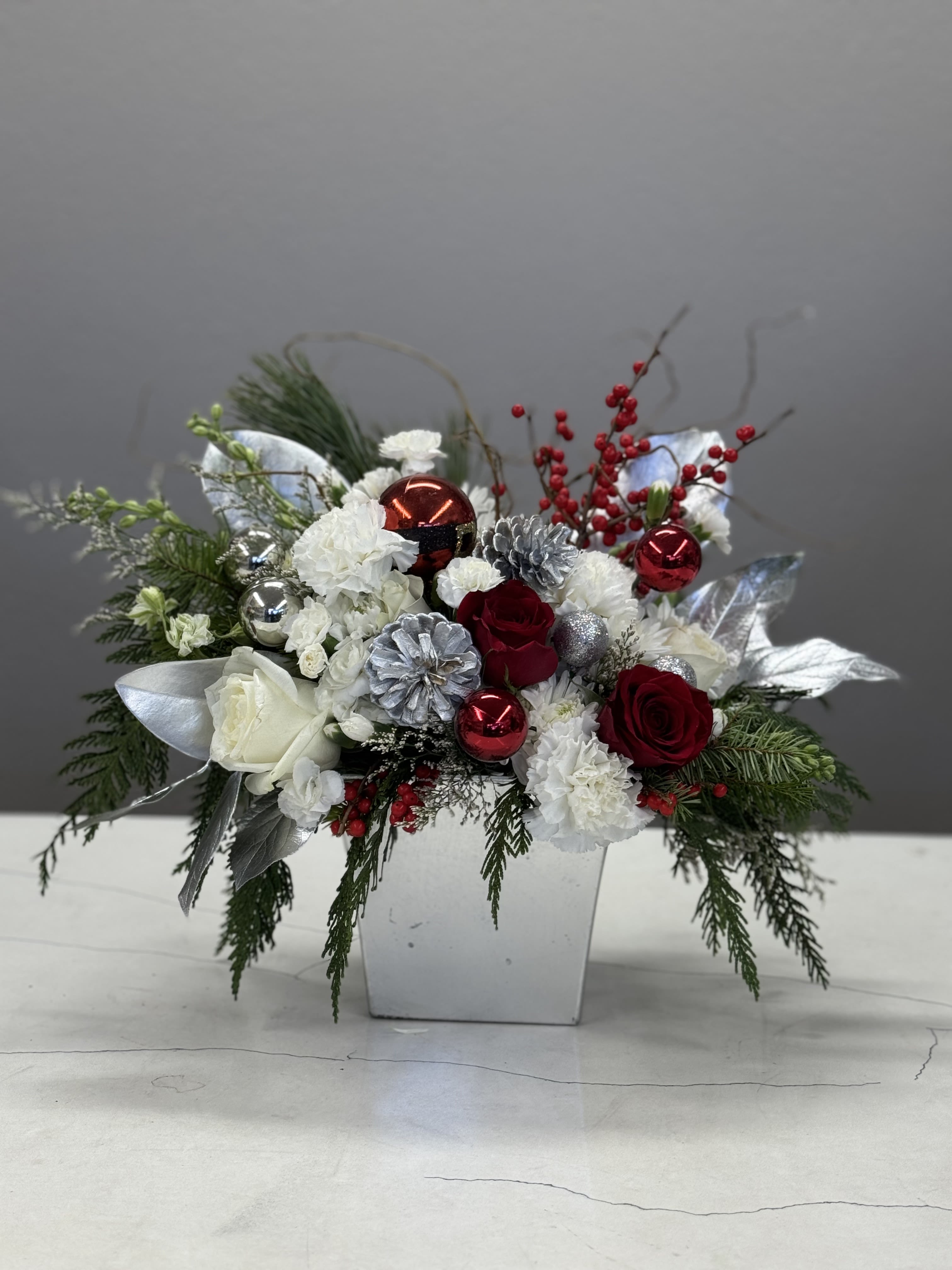 Crimson Frost - "Crimson Frost" is a striking and elegant floral arrangement that combines the timeless beauty of white, red, and silver. The rich red hues of luxurious roses and red berries bring warmth and passion to the design, contrasting beautifully with the pure, crisp white roses and carnations.  Silver foliages and pine cones intertwine through the arrangement, bringing a cool, frosty touch that highlights the vibrancy of the red and the softness of the white. The silver accents shimmer like a winter frost, elevating the entire composition and giving it an ethereal, almost magical quality.  Perfect for festive occasions, romantic celebrations, or holiday events, "Crimson Frost" captures the essence of winter's beauty—where fiery red meets serene white, all set against a backdrop of glistening silver.