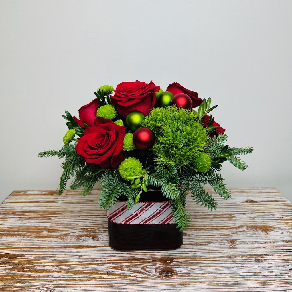 Holly Jolly - ‘Tis the Season to be Jolly! Our Holly Jolly is a very Merry and Bright arrangement of traditional shades of red and green flowers and ornaments in a red cubed vase accented with candy cane ribbon.   Approximately 10” Tall