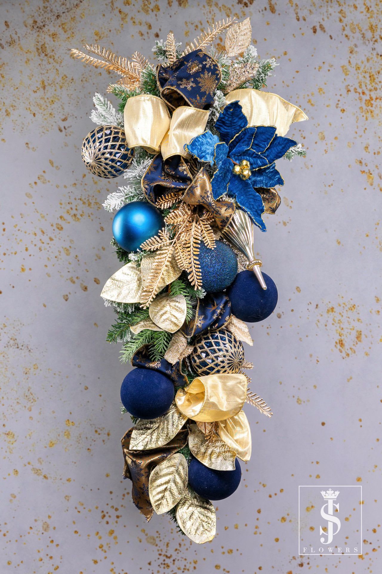Royal Blue Swag - The Royal Blue Swag is a regal holiday decoration that combines the elegance of deep sapphire blue with the opulence of shimmering gold. Featuring frosted greenery, golden leaves, and luxurious velvet blue ornaments, this piece exudes sophistication and festive charm. Embellished with intricate ribbons, glittering accents, and a statement blue bloom, it creates a stunning focal point for your holiday décor. Perfect for adorning doors, walls, or mantels, this handcrafted swag is a celebration of timeless beauty and holiday grandeur. Elevate your space this season with its rich, royal allure!