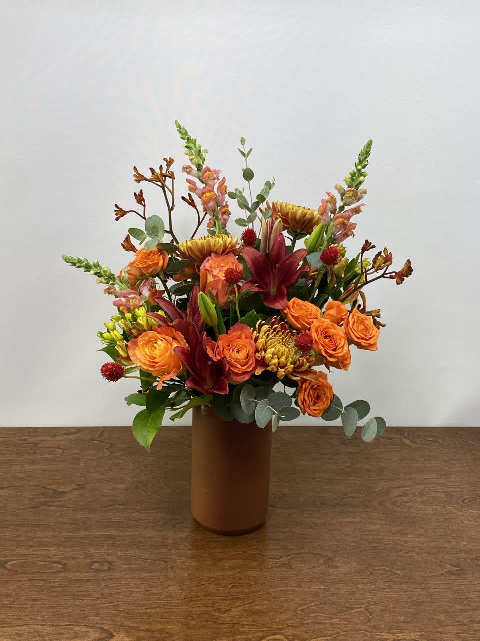 Autumn Garden - An arrangement of rose, lily, chrysanthemum, snapdragon, gomphrena, and kangaroo paw in warm fall colors. Arranged in a mahogany brown cylinder vase. This seasonal selection would be perfect as a gift for any fall occasion. 