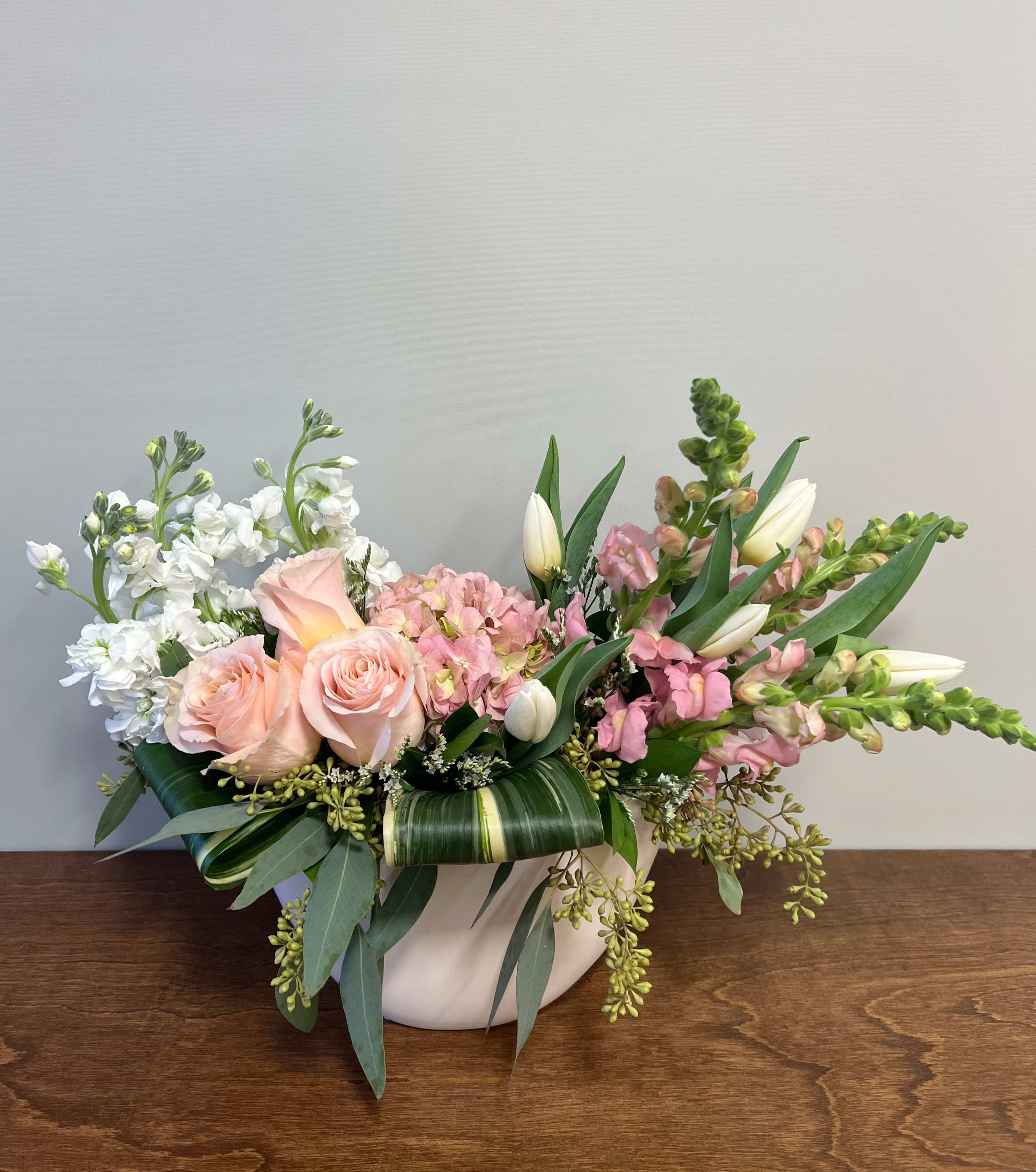 Crescent  - A graceful arrangement of soft colors in a crescent-shaped ceramic vase (8" by 5"). Flowers in this arrangement include tulips, roses, snapdragons, and stock. Please note, tulips and seeded eucalyptus are not in season year-round and will be substituted with the most similar flower or green available when out of season.  All versions of this arrangement come in the same vase. The deluxe and premium versions include either a higher number of flowers or higher value flowers.