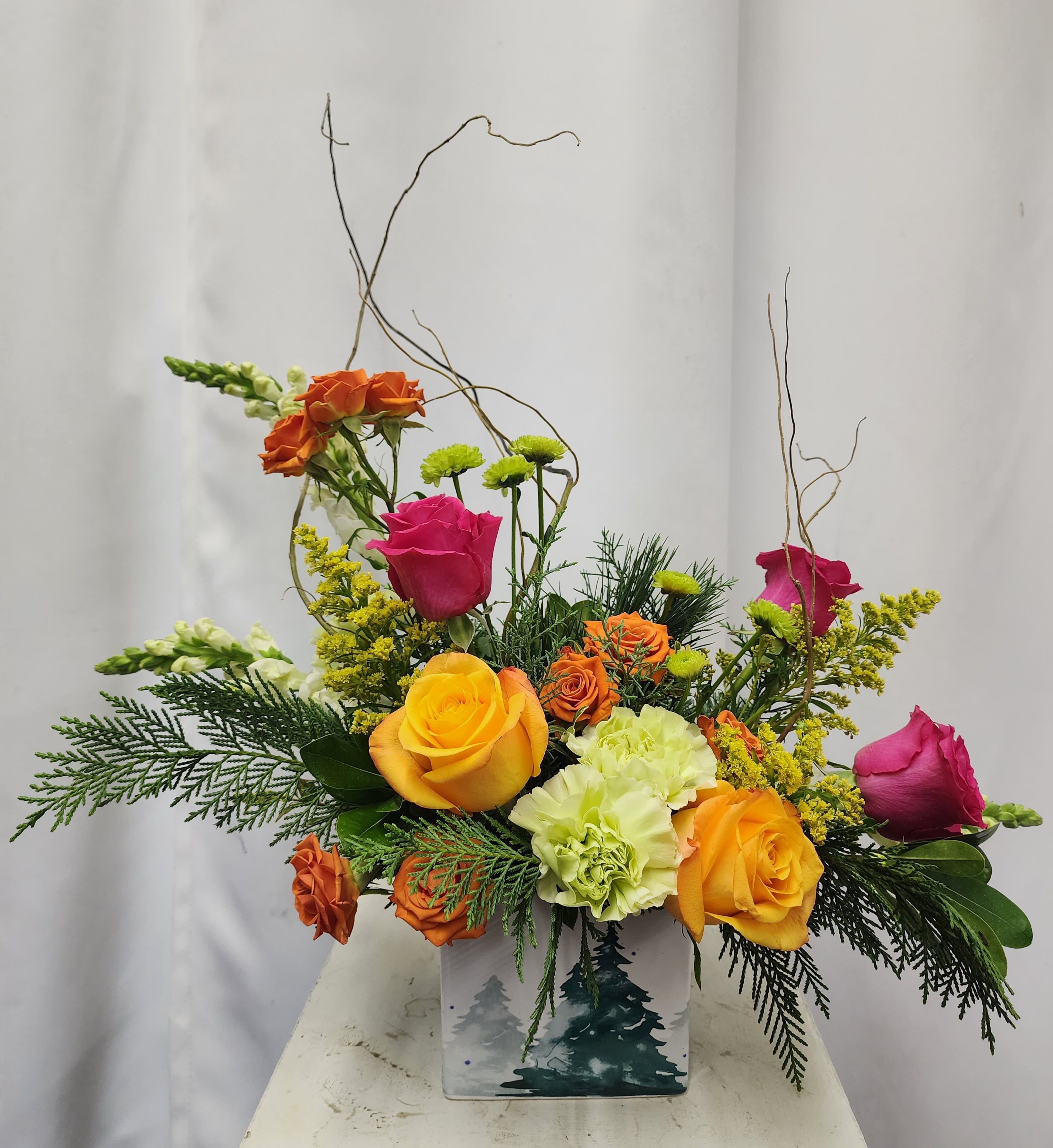 Vibrant Holiday Joy - Brighten any space with hot pink and vibrant orange roses, orange spray roses, green button poms, green carnations, and yellow solidago, all nestled in fresh holiday greens. Accented with curly willow and presented in a white ceramic cube keepsake featuring green trees. This arrangement brings a lively burst of color and festive charm to your holiday celebrations.