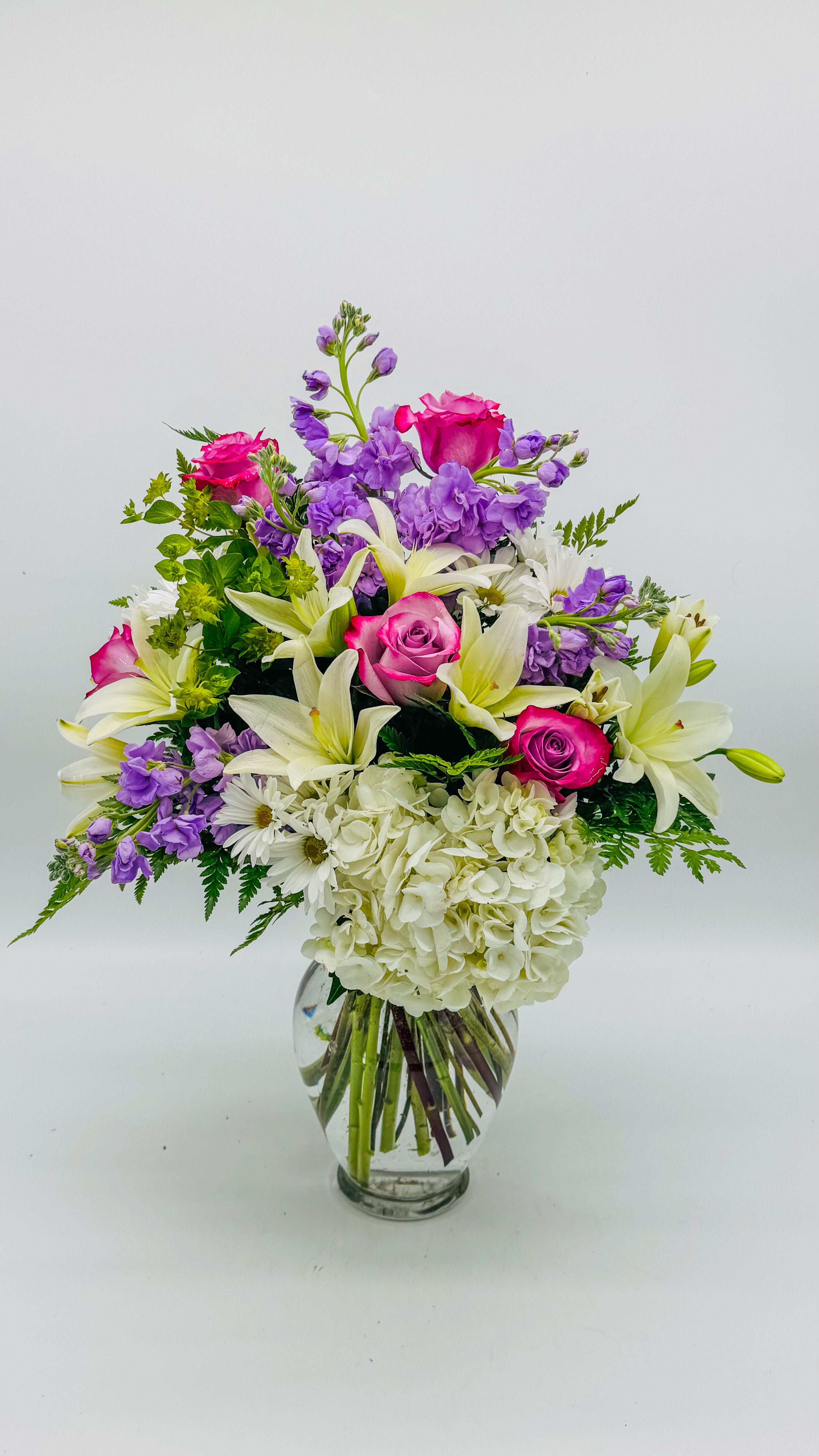 Lavender Sunset Symphony - A timeless celebration of elegance, this bouquet blends lavender and lilac tones with blush pink roses, ivory lilies, and soft white hydrangea. Delicate greenery and sprays of purple statice add depth, while the clear glass vase provides a clean, modern finish. Fresh, fragrant, and perfectly balanced, it’s ideal for anniversaries, celebrations, or simply brightening someone’s 