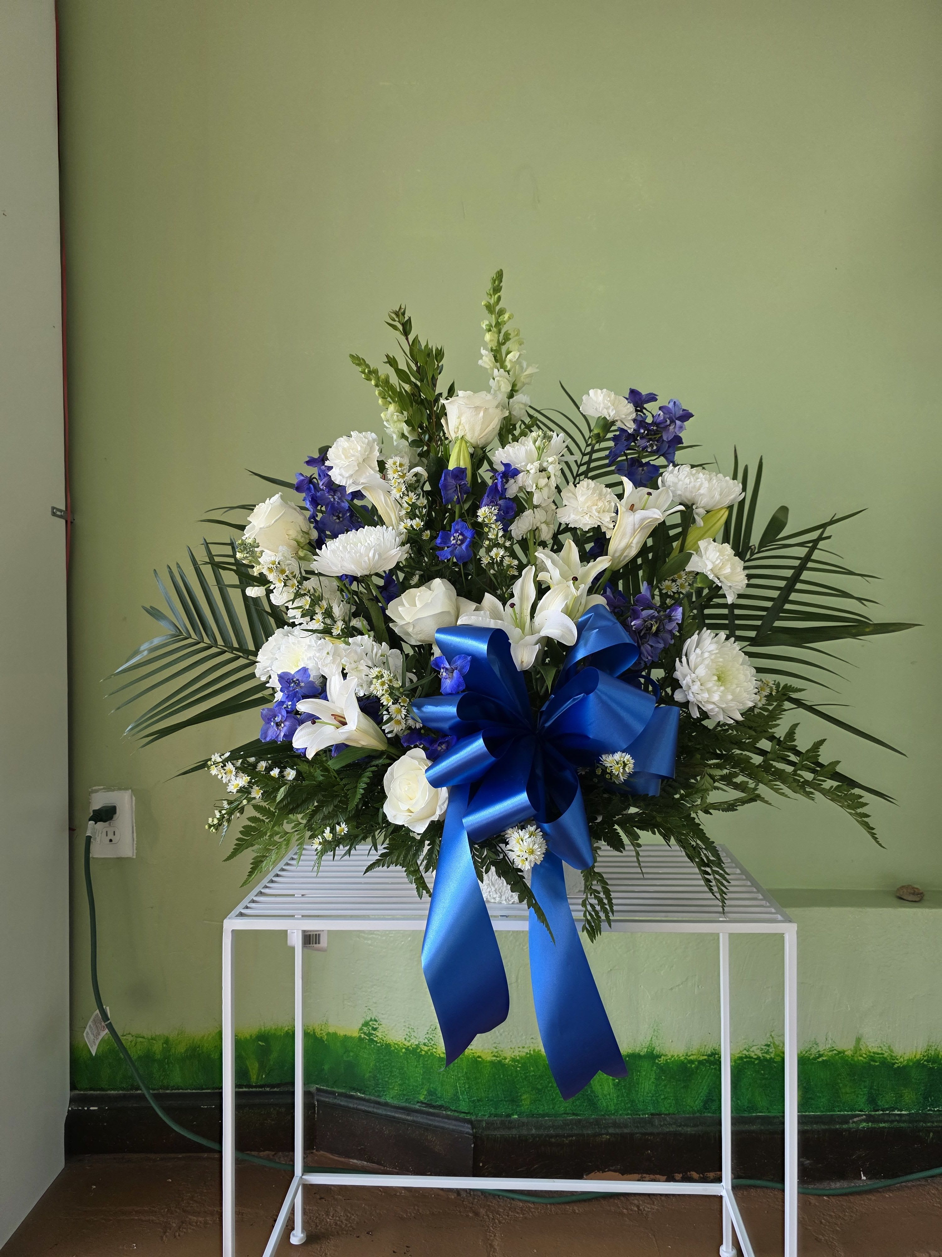 Peaceful Tribute - Elegant and serene, the Peaceful Tribute features a timeless blend of white roses, lilies, carnations, and chrysanthemums, symbolizing peace and remembrance. Accented with blue delphiniums and framed by lush greenery, this arrangement offers a heartfelt expression of sympathy.  Finished with a satin blue bow, it’s a beautiful tribute for funerals, memorials, or celebration of life services.