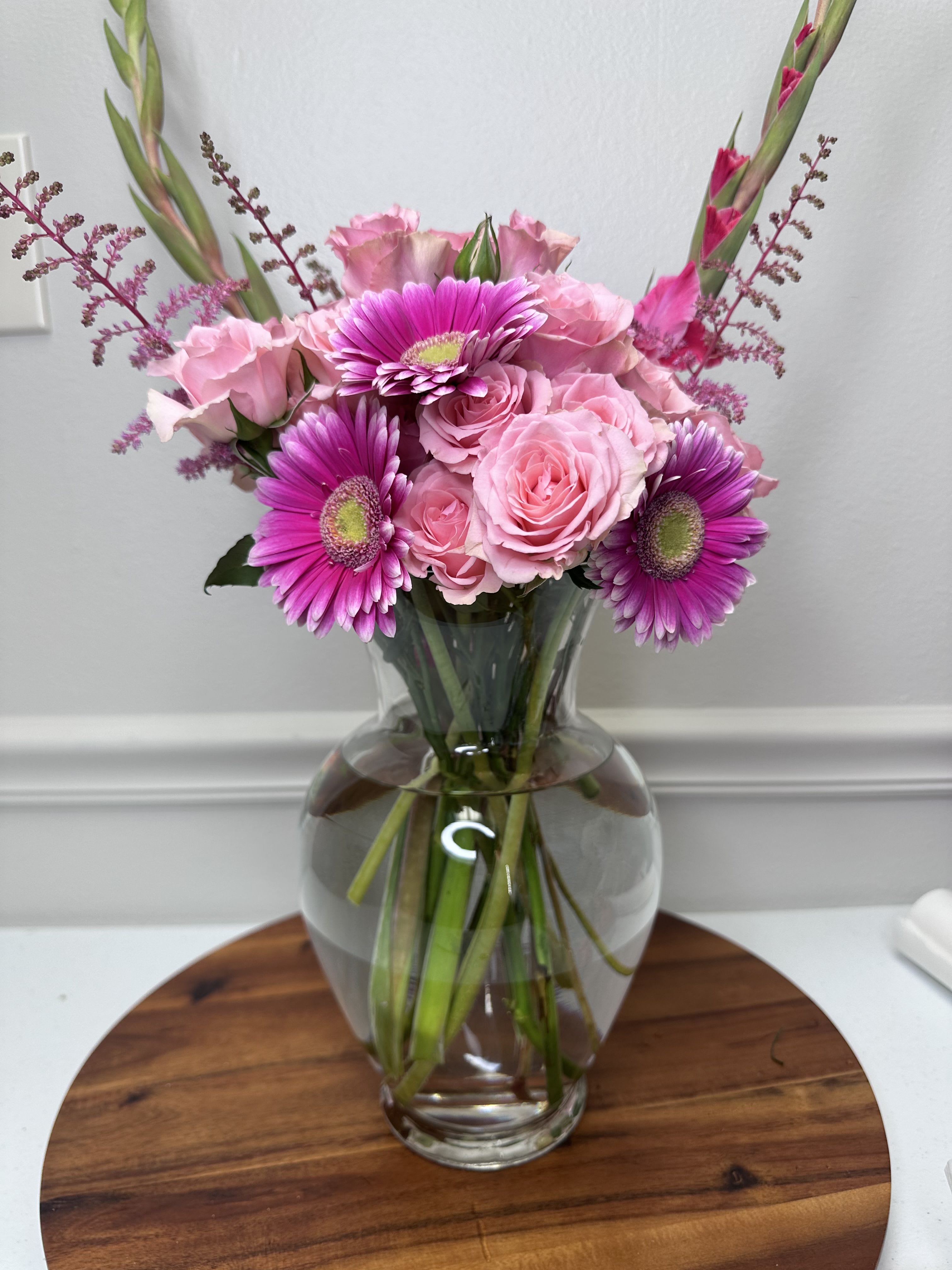 Rosy Dreams - A delicate and romantic arrangement presented in a glass vase, featuring soft pink roses, lilac gerberas, graceful gladiolus stems, and fine foliage accents that add height and lightness. The harmony of pink and lilac tones creates a feminine, sweet, and sophisticated design—perfect for brightening any space and expressing affection, admiration, and serenity.