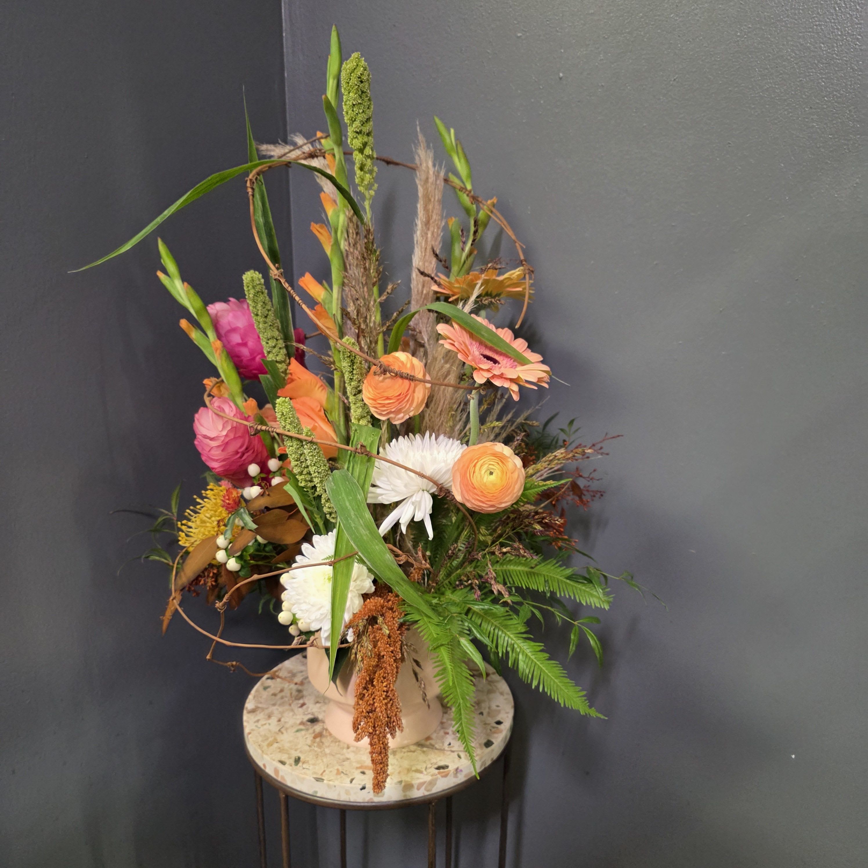 Fiery Pumpkin Spice  - Introducing our Fiery Pumpkin Spice  — a tall, sculptural fall arrangement bursting with rich seasonal color and wild, textural movement. Designed in a modern palette of burnt orange, coral, golden yellow, fuchsia, and creamy white, this piece features ranunculus, gerbera daisies, chrysanthemums, gladiolus, pampas grass, millet, fern, and autumn foliage for a dramatic harvest-inspired look.  Earthy grasses and twisting branches create height and organic shape, while bold blooms add vibrant pops of color reminiscent of a Colorado fall sunset. This eye-catching design is perfect for entry tables, event décor, corporate gifting, and luxury flower delivery across Denver, Centennial, Cherry Creek, and Greenwood Village.  A standout fall piece for anyone who loves color, texture, and modern botanical design.