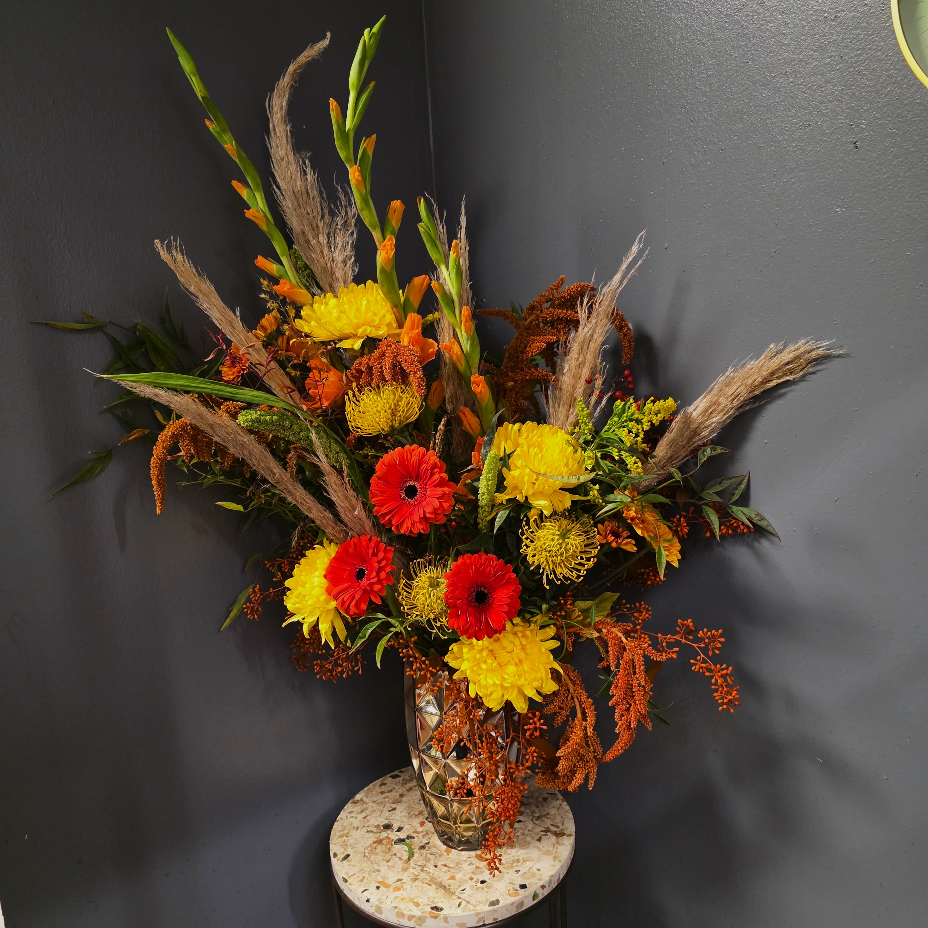 Harvest Showstopper - Introducing Harvest Showstopper, a dramatic autumn statement arrangement designed for those who love bold color, texture, and luxury. Created in a faceted gold vase, this towering fall piece explodes with sunset-hued gerbera daisies, golden and tangerine mums, orange gladiolus, pincushion protea, lush fall foliage, and rust-colored amaranthus, all accented with soft pampas plumes for height and movement.  This high-impact design evokes the warmth of a Colorado fall sunset — fiery reds, glowing golds, and rich amber tones layered with wild, organic textures. Perfect for entryways, hotel lobbies, holiday gatherings, corporate gifting, or Thanksgiving centerpieces, this arrangement brings a luxurious seasonal presence to any space.  Available for same-day flower delivery in Denver, Centennial, Cherry Creek, Greenwood Village, and surrounding areas, this oversized fall bouquet is ideal for clients who want a truly spectacular autumn floral experience.