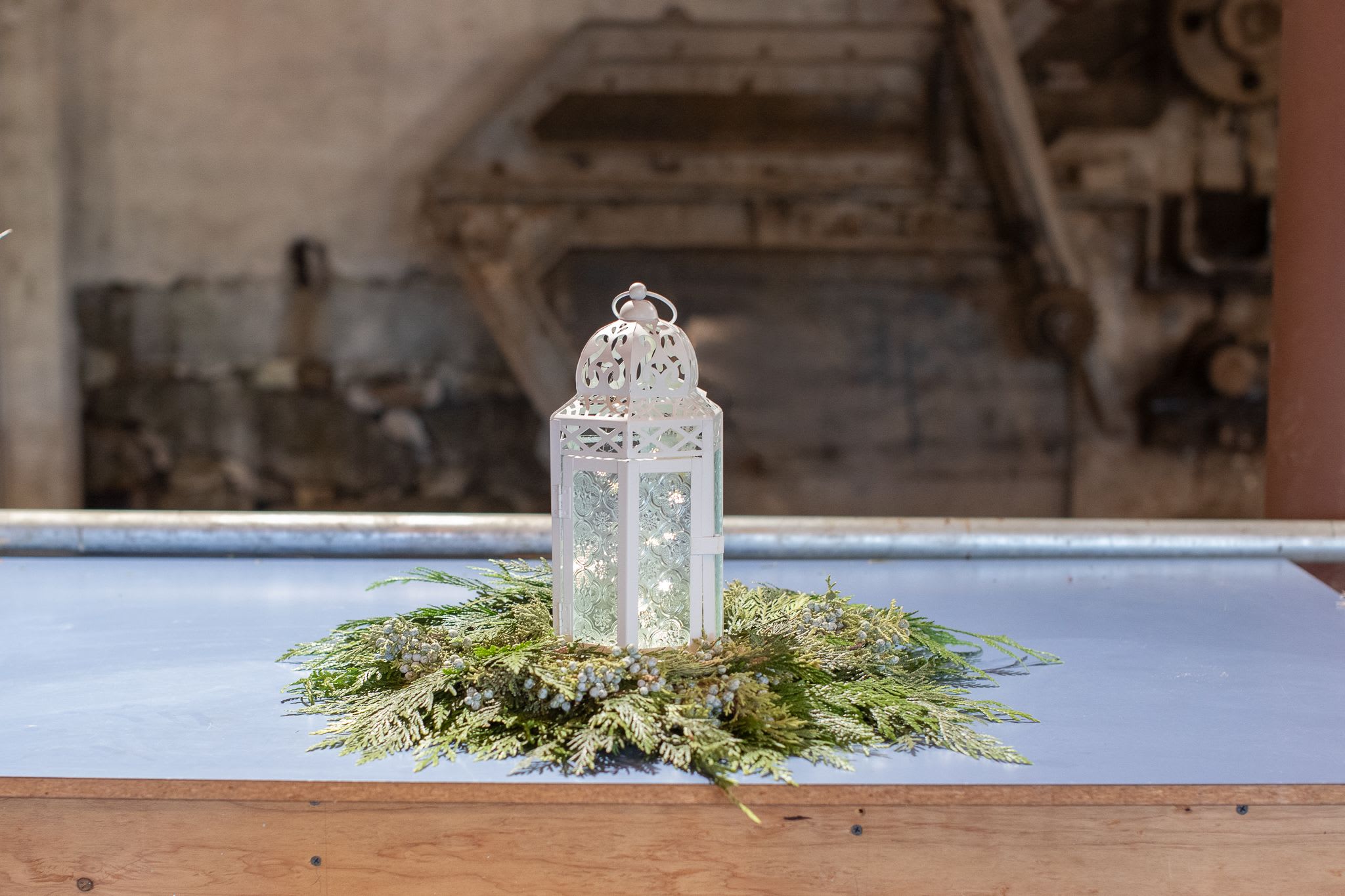 Cedar Simplicity Centerpiece  - A simple a beautiful long lasting centerpiece that awakens the senses. Fragrant cedar and juniper are woven into a ring that can brings seasonal scent into the home. This centerpiece is perfect for gifting in winter. A white embossed lantern is placed in the center with glowing fairy lights. The cedar and juniper ring will eventually dry preserving itself. Minimal care and assembly required for the recipient. The wreath of cedar spans about 12-18 inches at its widest point(s) in a circular fashion. The lantern is about 12 inches in height. The centerpiece will be delivered on the flat piece of cardstock, we suggest leaving it as a barrier between with fresh foliage and wood surfaces. This can be used in so many ways as a decorative accent piece. The lantern is a wonderful keepsake gift. Call with any questions or stop by and pick one up today. Limited quantity.   