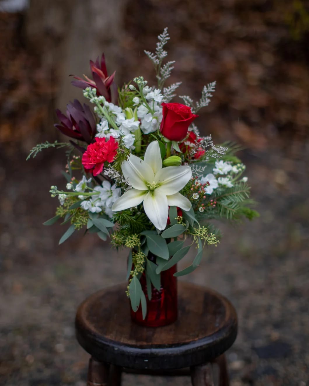 Christmas Peace Bouquet - Pretty white lily and red roses with carnations nested on bountiful winter foliage in a ruby red textured festive vase with an embossed poinsettia flower printed in the center on either side. Limited supply of red vases, once they are sold out clear glass will be utilized.