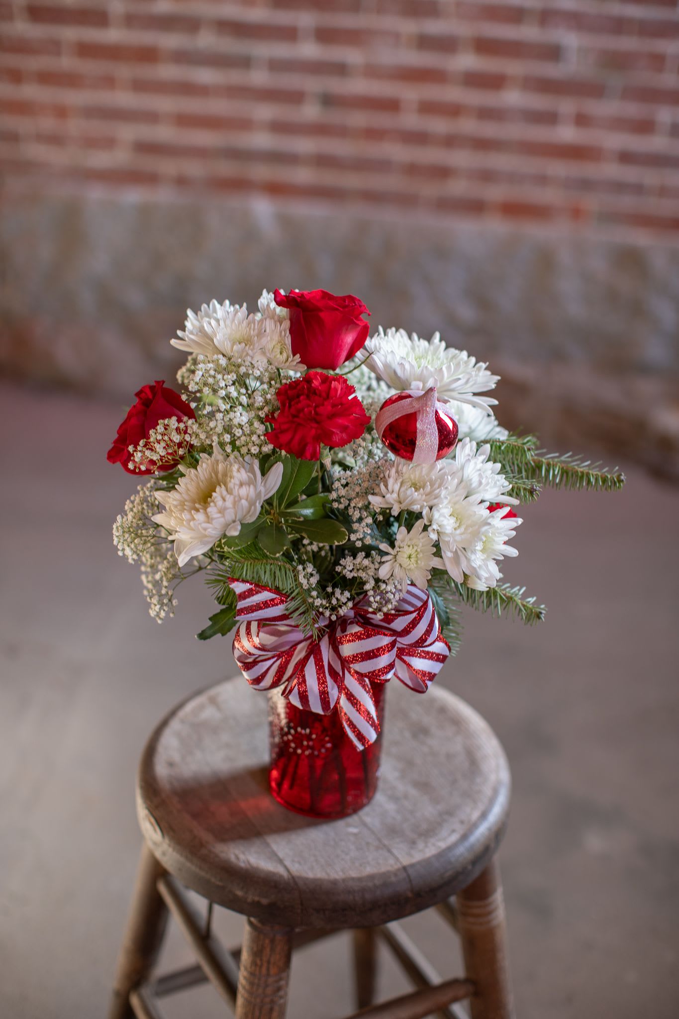 Peppermint Bliss - A ruby red textured jar vase holds crisp, sweet and spicy blooms. This design is complete with a festive decorative blown glass ornament charm of red and white. Simple foliage of pittosporum and pine give the arrangement winter feels. Floral varieties include cremons, cushion pom pons, red carnations and a rose. Accent flower may include gypsophila more commonly known as babies breath. This novelty design is finished with a tidy manicured bow of peppermint striped ribbon of red and white. Oriinal design by Katie.