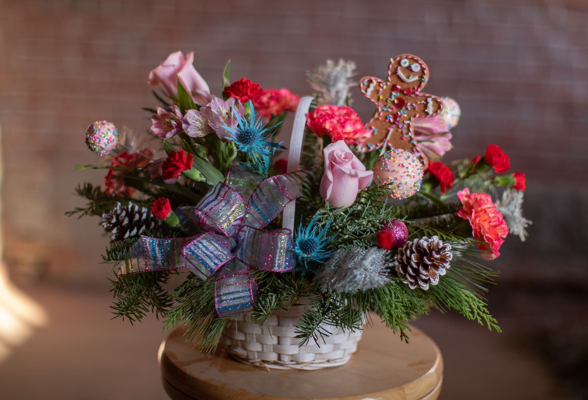 Bakery Gingerbread - &quot;You can't catch me, I'm the gingerbread man!&quot; This whimsical design of winter delights is created in a woven wicker basket featuring an adorable gingerbread man decorative pick. Artificial decorative  sweets with pastel ornament decorations are neatly arranged in a two sided style. Balsam, Carolina sapphire, pine and more are a few of the greenery selections featured. Fun-loving assorted carnations chase the thistle and roses as fast as the can in this white wash basket complete with a sparkling colorful striped handcrafter bow. Original design by Katie.