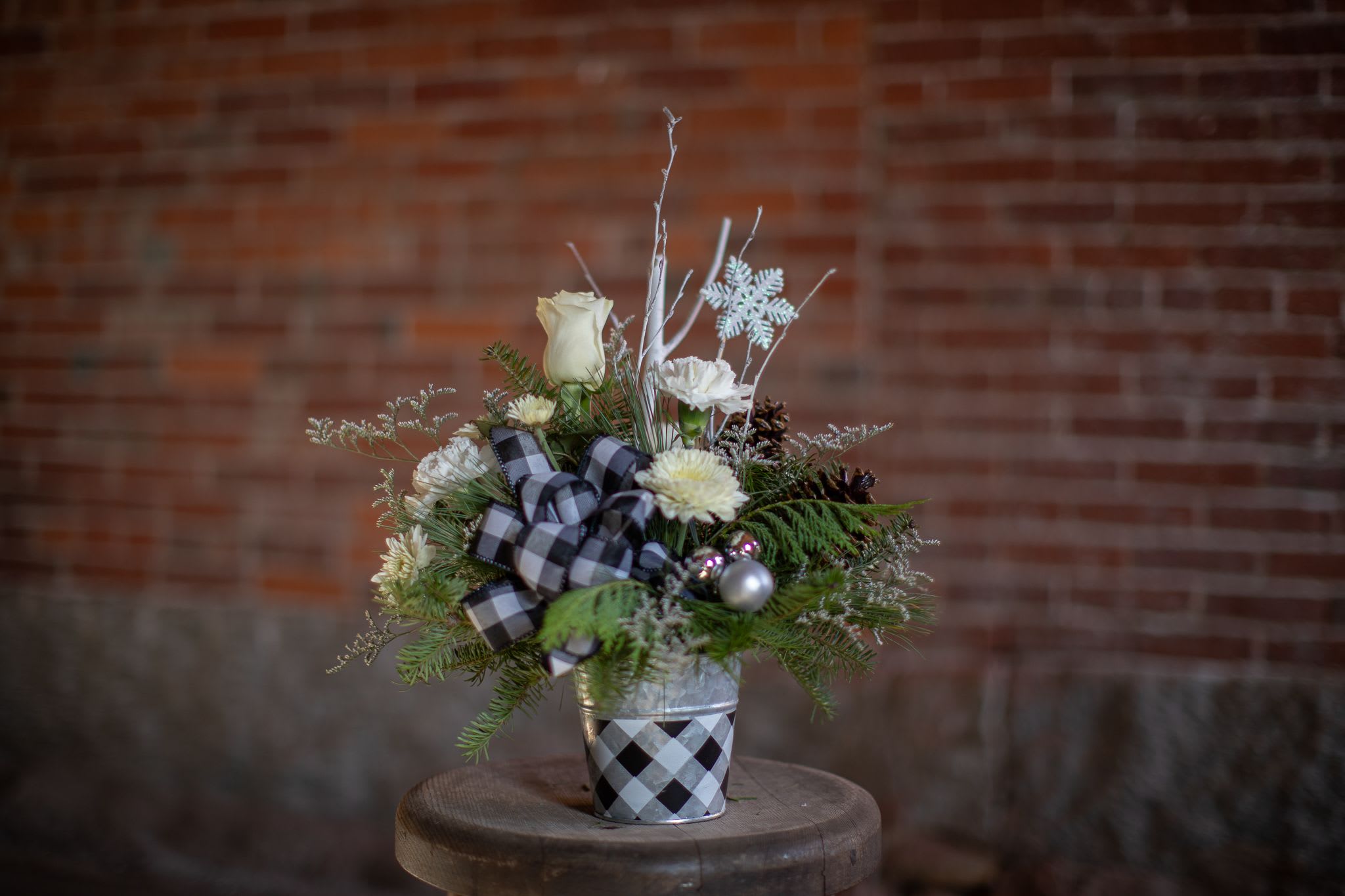 Winter Farmhouse - A winter buffalo plaid themed arrangement for the season. A printed tin with a black and white buffalo checked pattern. Simple white branches and flowers are carefully designed around fragrant foliage varieties of cedar, pine and balsam. Assorted silver ornaments, lacquered pinecones and an iridescent snowflake pick are unique design elements included in this arrangement. A buffalo plaid handcrafted bow complete the winter farmhouse look. Standard size is created in a 4 inch tin container. Deluxe and premium size in 6 inch size tine container.