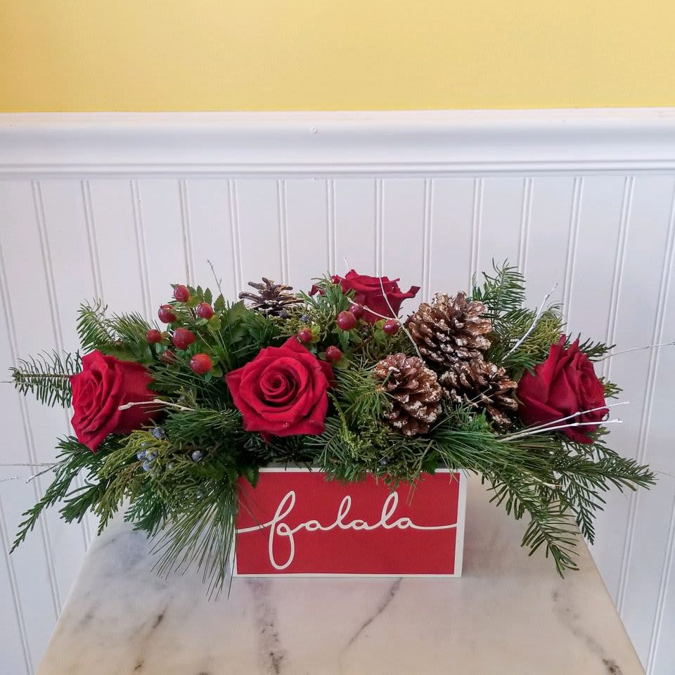 Festive Fa La La  - This arrangement features vibrant red roses, lush pine branches, pinecones, and holly berries, all elegantly arranged in a rectangular red container with &quot;falala&quot; written in white cursive on the front. It's the perfect holiday centerpiece to bring cheer and warmth to any space. 