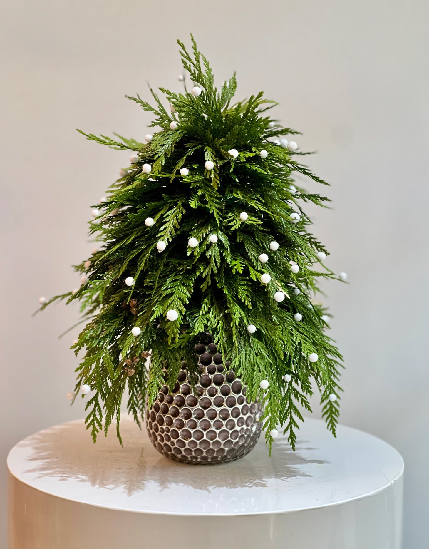 Winter Evergreen Mini Tree - Bring the spirit of the holidays into any space with this chic, modern mini tree designed entirely from fresh cedar. Styled with a soft, natural drape and adorned with delicate white berry accents, this piece captures the magic of winter in an elegant, minimalist form.  Set in a designer textured vase, the arrangement blends organic beauty with contemporary luxury—perfect for entryways, desktops, kitchen islands, or as a festive gift. Fresh, fragrant, and full of charm, this evergreen creation elevates holiday décor with effortless sophistication.