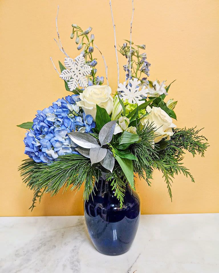 Winter Bliss Bouquet - This stunning floral bouquet is elegantly arranged in a dark blue vase, featuring a beautiful mix of white roses, blue hydrangeas, and lush greenery, including pine branches and silver leaves. Decorative white snowflakes and delicate white branches add a festive, wintery touch, making it the perfect centerpiece for any holiday celebration or winter decor. 