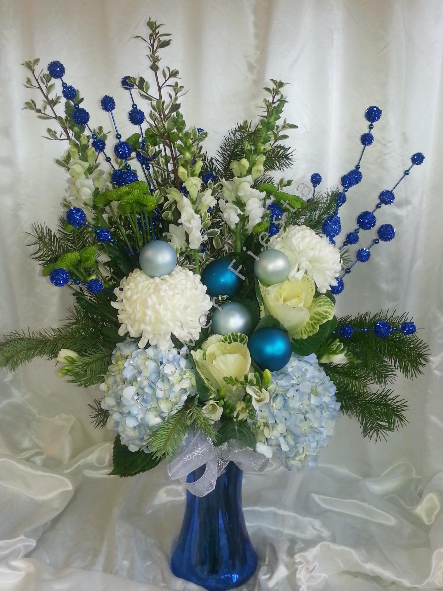 Hanukkah Arrangement - Beautiful cool shades of white and blue alongside ornaments for the holiday and winter greens in a royal blue gathering vase.  Order well in advance. Items may vary.  