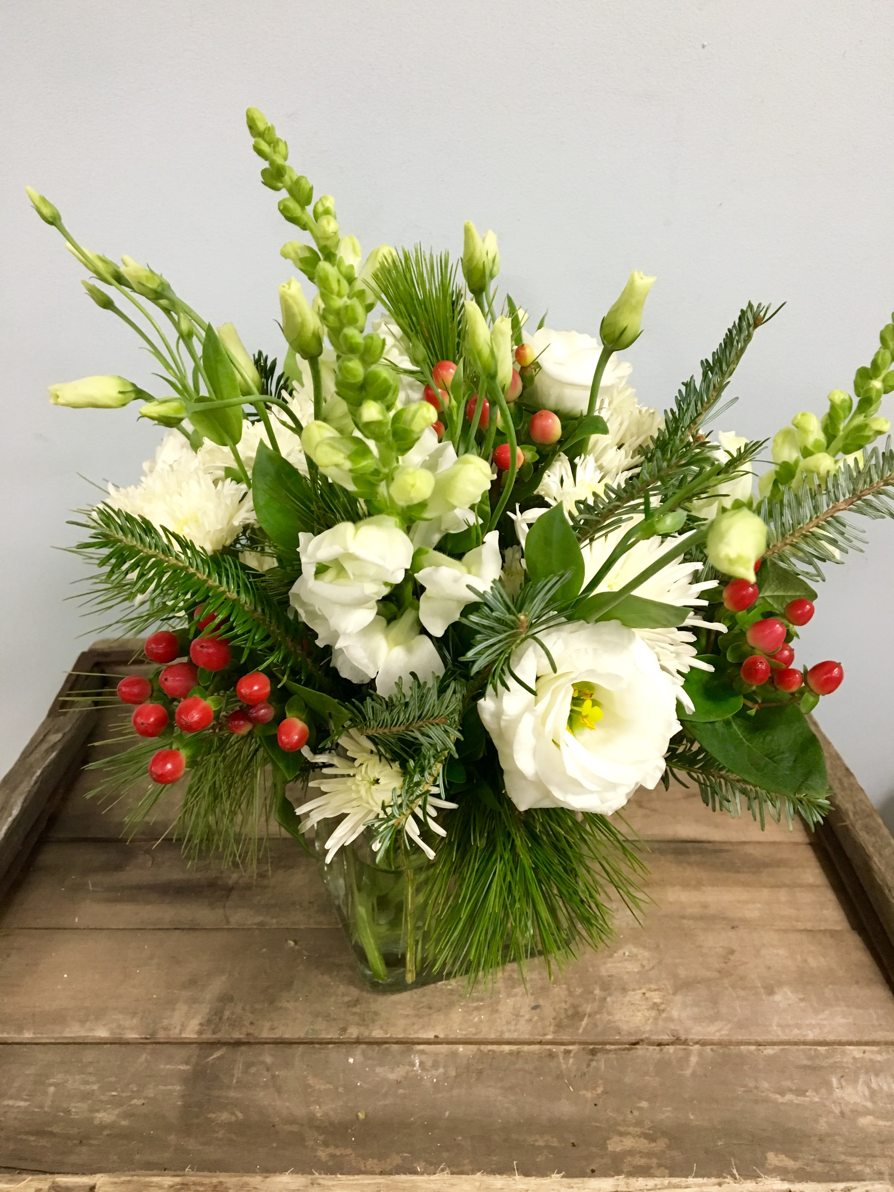 Winter Berry Forest - Simple and seasonal, this arrangement will make a great centerpiece or gift for someone's desk. Seasonal white flowers are accented with red berries and designed in a glass cube bursting with the aroma of classic winter pine and balsam greenery. 