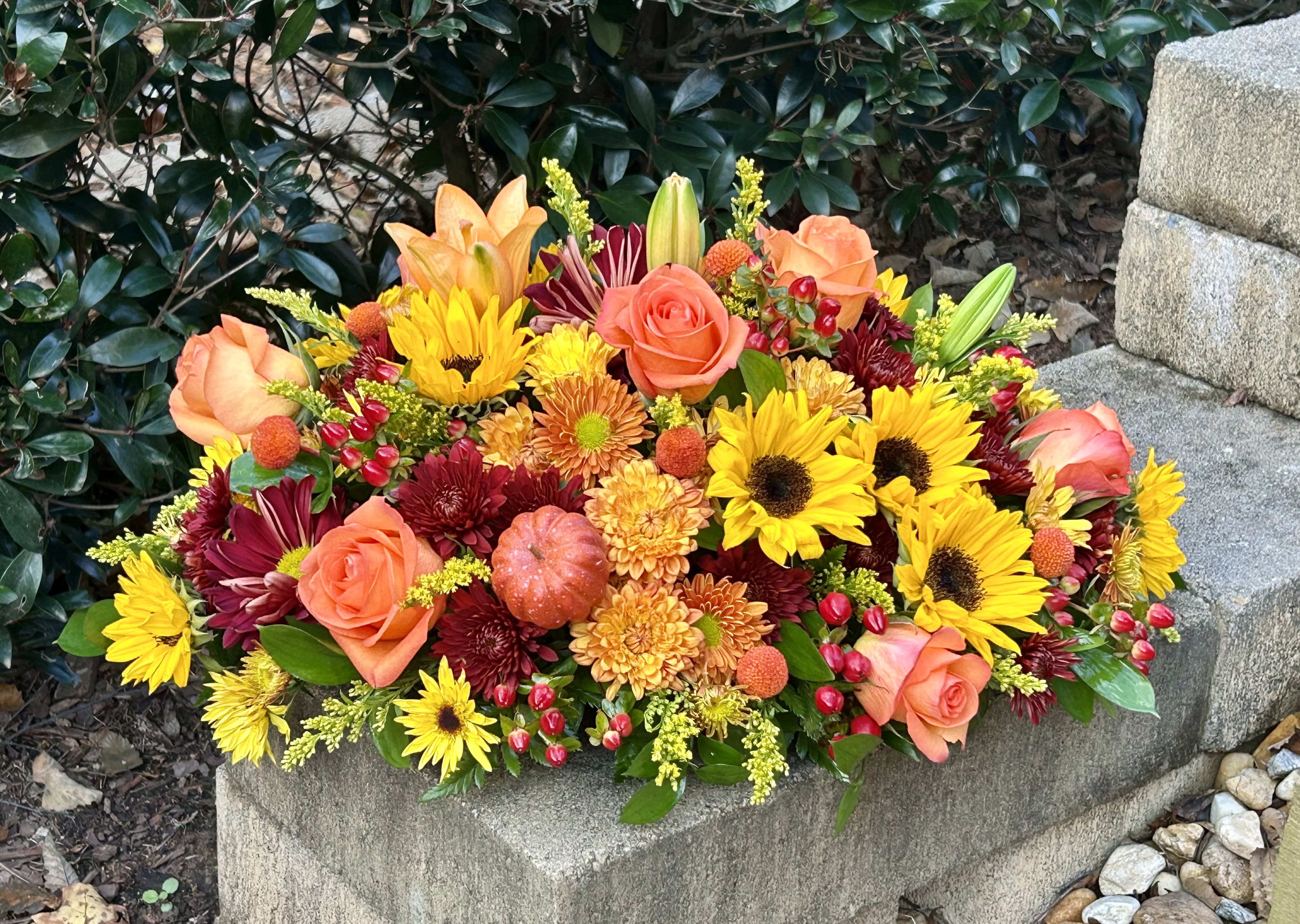 Autumn Harvest Glow Centerpiece - A warm, vibrant fall centerpiece bursting with golden sunflowers, orange roses, burgundy mums, and playful touches of mini pumpkins and berries. Arranged in a lush, low profile, it brings the rich colors and cozy spirit of autumn to any table or gathering.