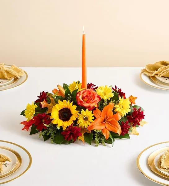 Fields Of Europe Centerpiece - The rich beauty of the European countryside comes alive in our best-selling fall centerpiece. Crafted by our artisan florists, it’s filled with a gathering of fresh-picked blooms in brilliant colors and finished with an orange candle to warm hearts and homes. • All-around centerpiece arrangement with autumn-colored roses, sunflowers, red daisy poms, orange Asiatic lilies, mini carnations and solidago; accented with assorted greenery and dried oak leaves • Includes a 12&quot;&quot;H orange taper candle (overall dimensions do not include candle)