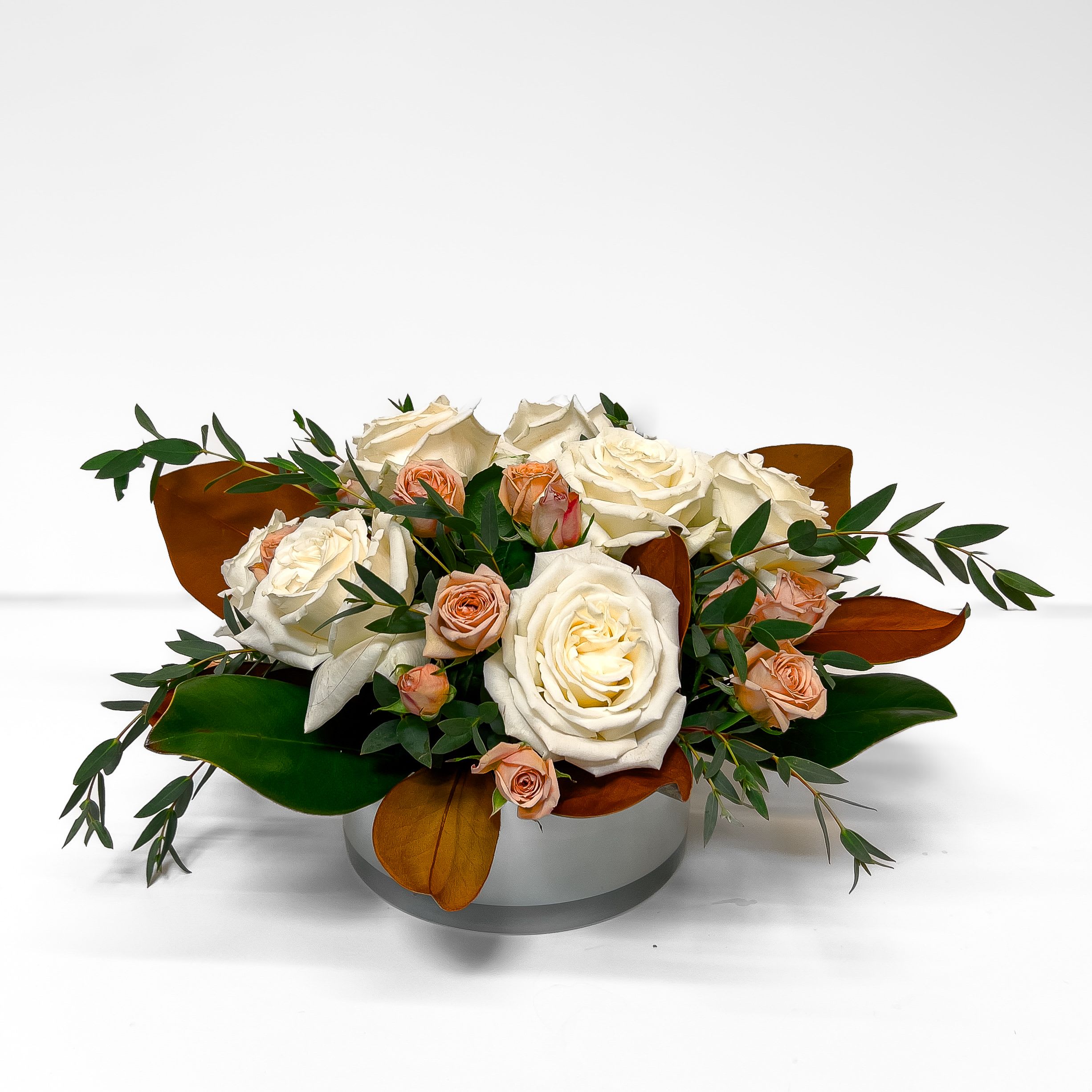 Magnolia Grace - White roses and magnolia leaves form a serene, low centerpiece that radiates quiet refinement. A breath of calm luxury for the autumn table. *PLEASE NOTE:  We have sold out of the white vase shown, we will be substituting with a leaf-lined, clear glass version of the same container.