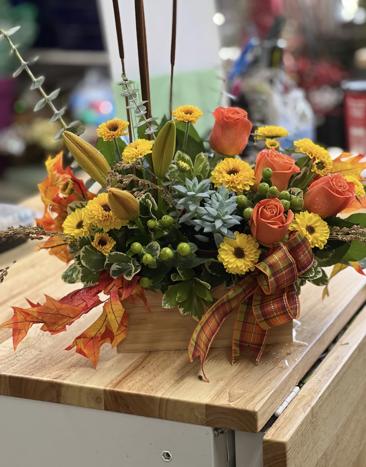 Autumn Harvest Box Arrangement - A refined seasonal design featuring vibrant orange roses, golden chrysanthemums, lilies, succulents, and natural greenery arranged in a wooden box for a warm, polished presentation. Accented with autumn leaves, hypericum berries, cattails, and a plaid harvest ribbon, this centerpiece brings a rich touch of fall elegance to any table or entryway. Ideal for home décor, holiday gatherings, or gifting during the autumn season.