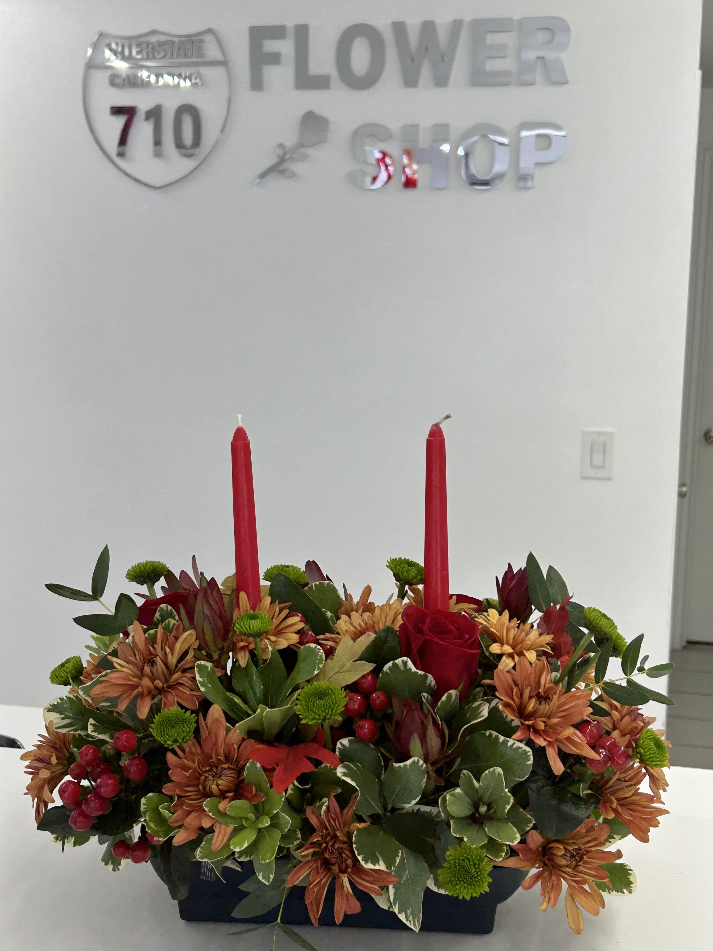 Golden Ember Centerpiece - A warm and festive Thanksgiving centerpiece filled with autumn-toned blooms, lush greenery, seasonal berries, and two classic red taper candles. Designed to bring cozy holiday charm to your dining table, this arrangement sets the perfect mood for gathering, gratitude, and celebration. Ideal for Thanksgiving dinners, holiday hosting, or gifting. 