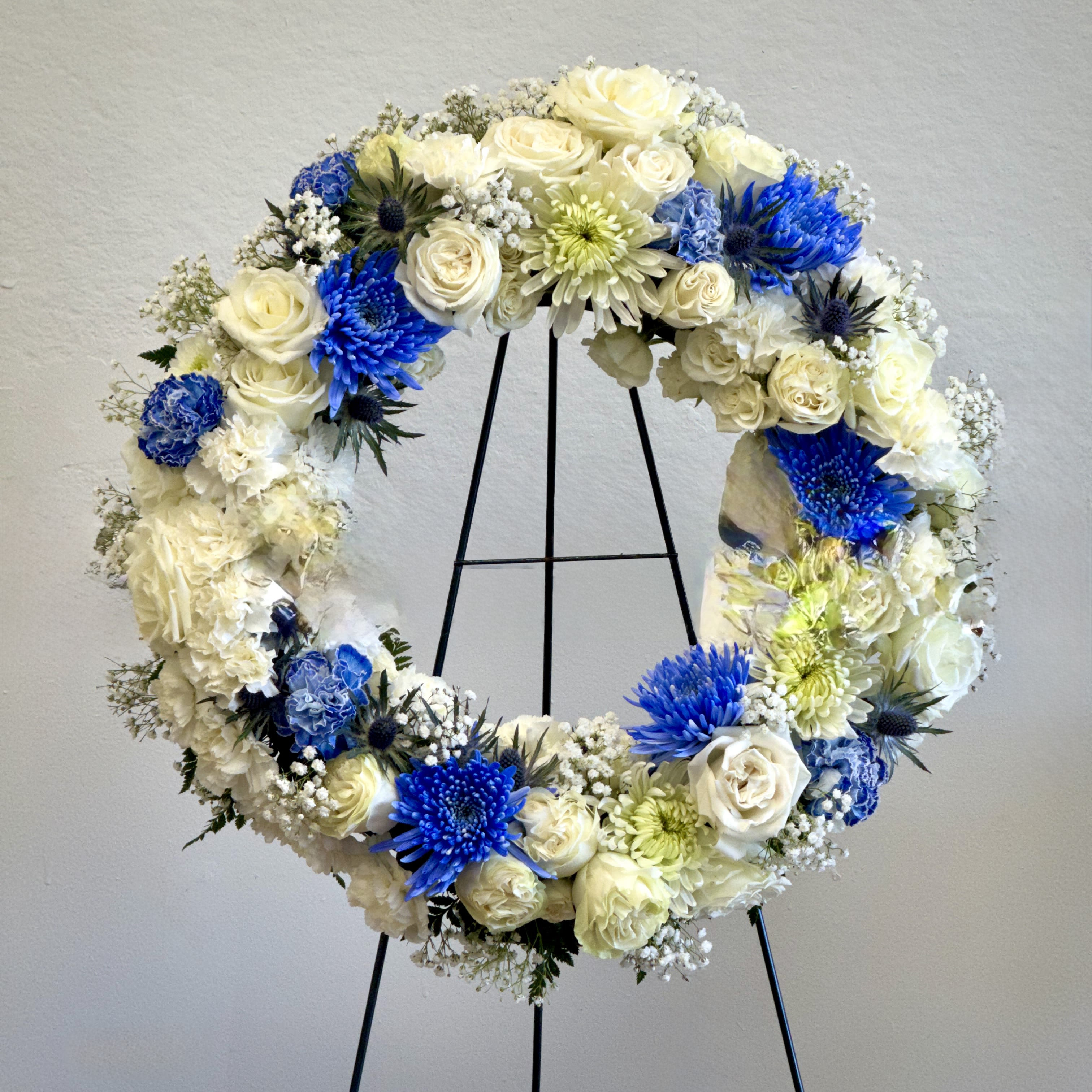 White and Blue Wreath II - A graceful sympathy wreath featuring a serene blend of white and royal blue blooms. Elegant roses, chrysanthemums, carnations, and accent florals are carefully arranged to symbolize peace, honor, and heartfelt remembrance. Displayed on a stand, this arrangement offers a beautiful tribute, conveying love and support with dignity and grace.