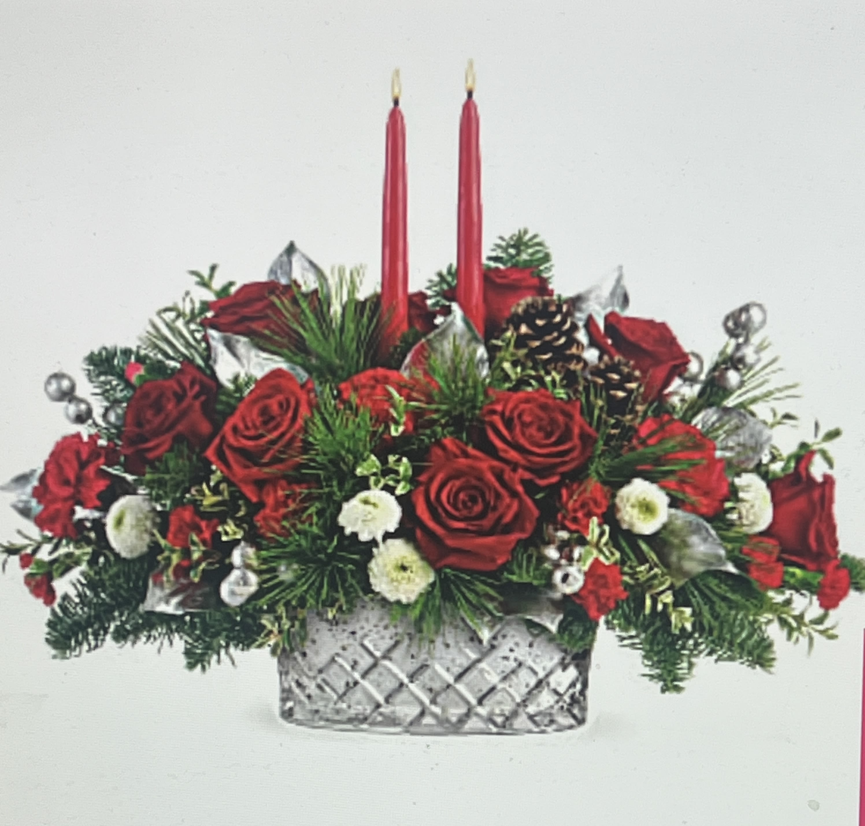 2025 Merry Christmas centerpieces by Charlene - Capture the magic of Christmas with this lush winter rose centerpiece, arranged with glowing tapers in a sparkling crosscut mercury glass bowl that will be a holiday treasure for years to come! Red roses, red carnations, red miniature carnations, and white button spray chrysanthemums are accented with noble fir, white pine, oregonia, lemon leaf, and red candles.*Reasonable flower substitutions may be made due to availability. 