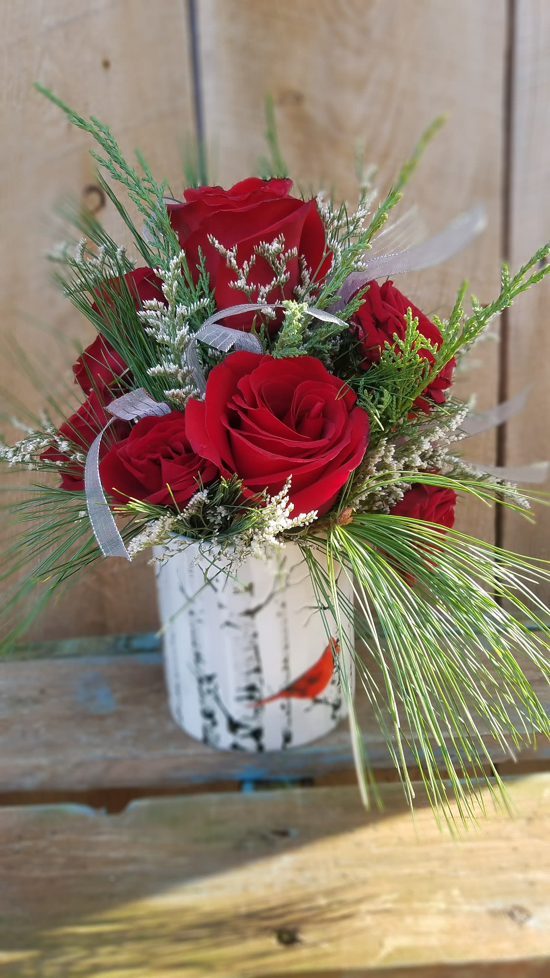 Through the Woods - A beautiful combination of white pine with red roses and silver ribbon in a white ceramic keepsake vase featuring a scene of a red cardinal in the woods. 